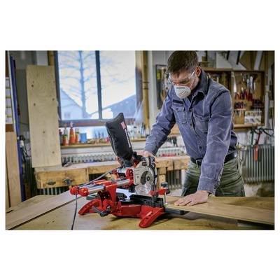 Un uomo che indossa una mascherina aziona una sega a troncatrice rossa in un laboratorio, tagliando una tavola di legno su un banco da lavoro.