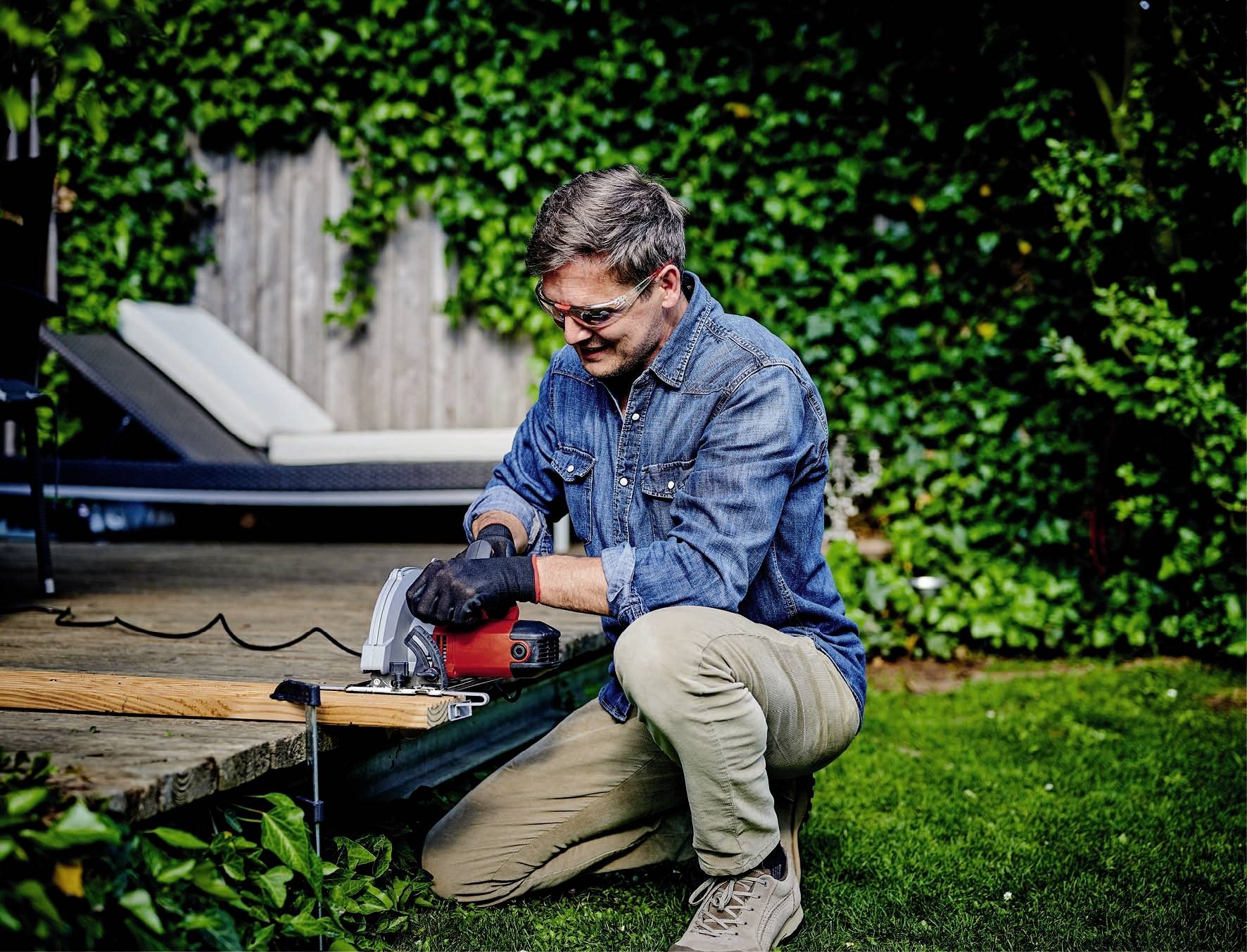 Un uomo in abiti da lavoro è inginocchiato in un giardino e taglia una tavola di legno con una sega elettrica. Sullo sfondo ci sono piante e un lettino.