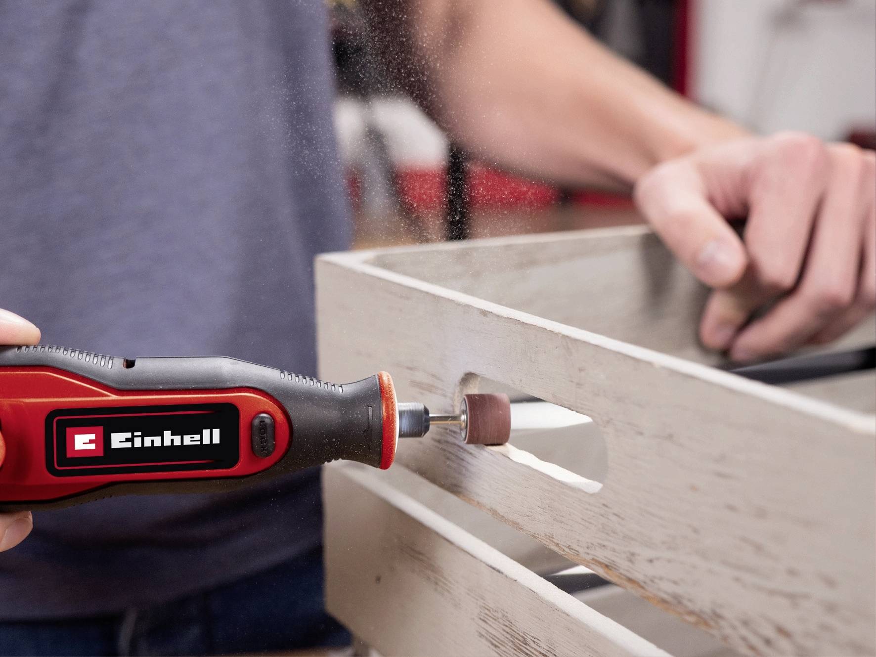 Una persona sta levigando un pezzo di legno con un utensile rosso Einhell. Trucioli di legno volano mentre la superficie viene lavorata.
