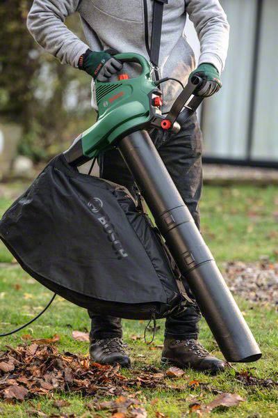Una persona sta utilizzando un soffiatore per raccogliere le foglie in giardino. Il soffiatore è verde e dotato di un contenitore di raccolta nero.