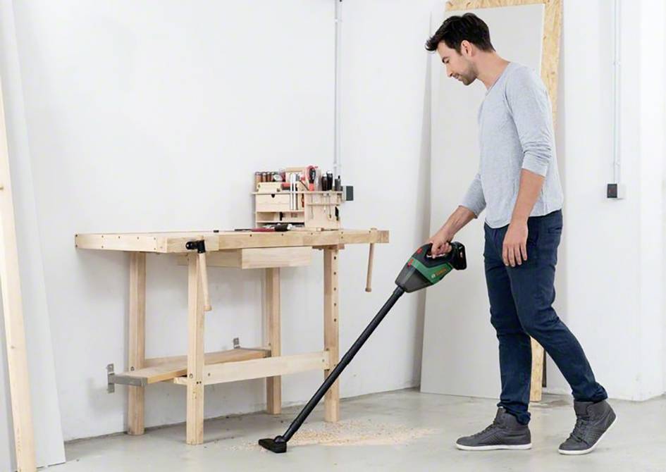 Un uomo sta in un laboratorio e aspira trucioli di legno dal pavimento con un aspirapolvere a batteria. Un banco da lavoro si trova sullo sfondo.