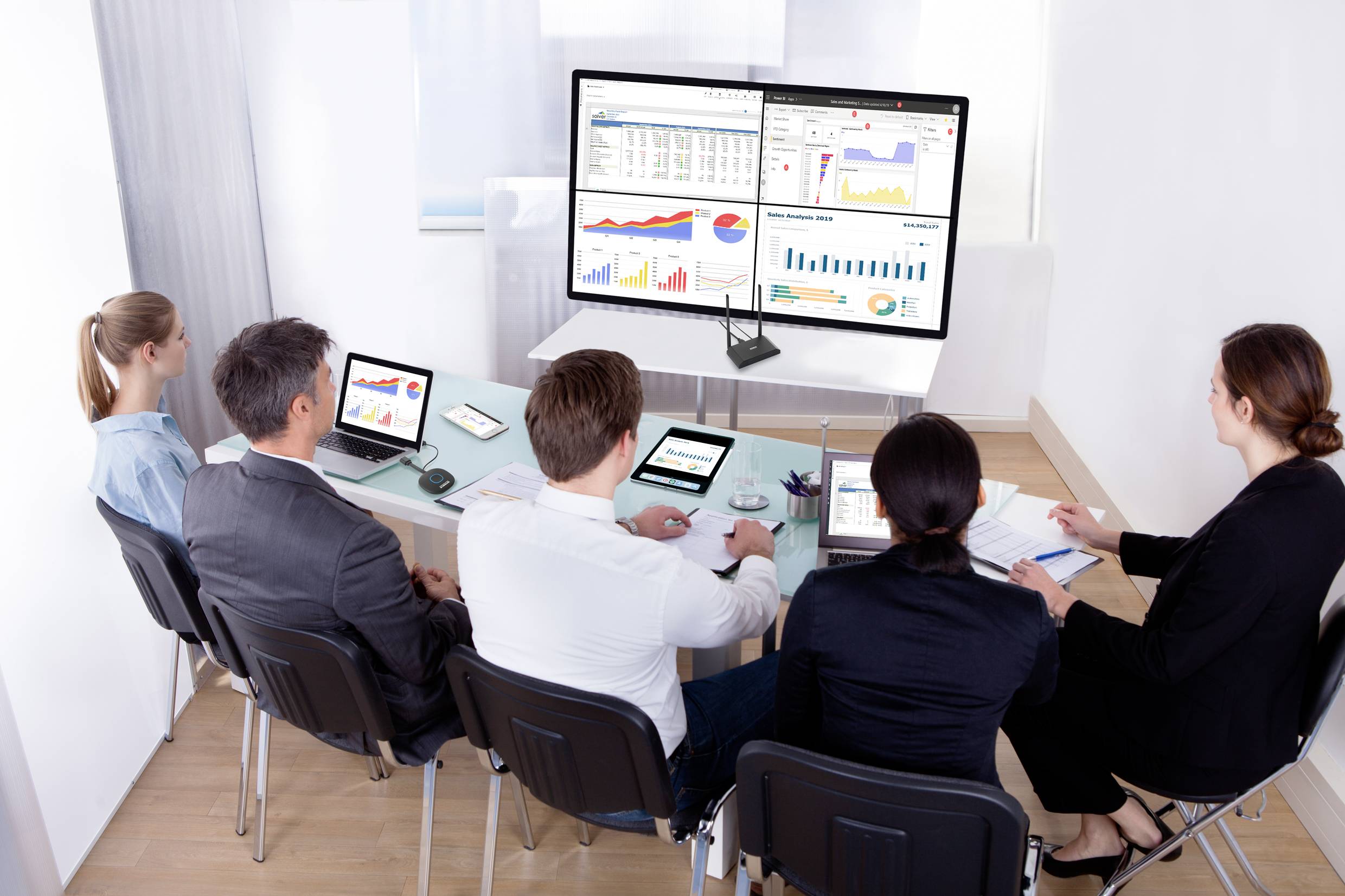 Un gruppo di cinque persone è seduto intorno a un tavolo in una sala riunioni, osservando una grande presentazione su schermo con diagrammi e grafici.