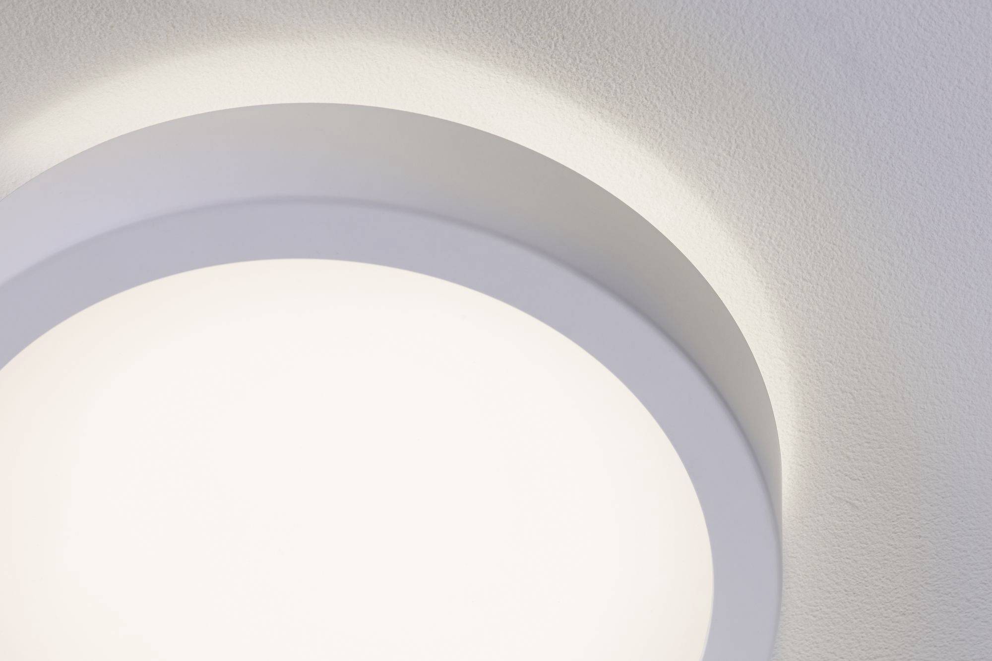 Una lampada da soffitto rotonda e bianca illumina intensamente una parete bianca. Effetti di ombra incorniciano la luce.