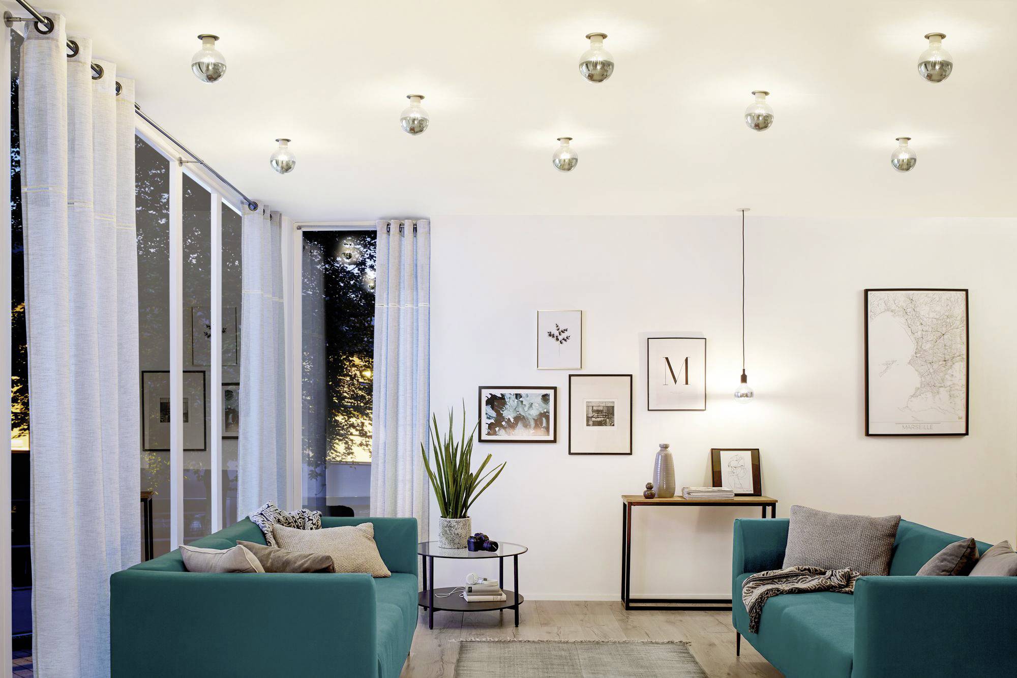 Soggiorno con pareti bianche, due divani turchesi, diverse lampade a soffitto e quadri decorativi alle pareti.