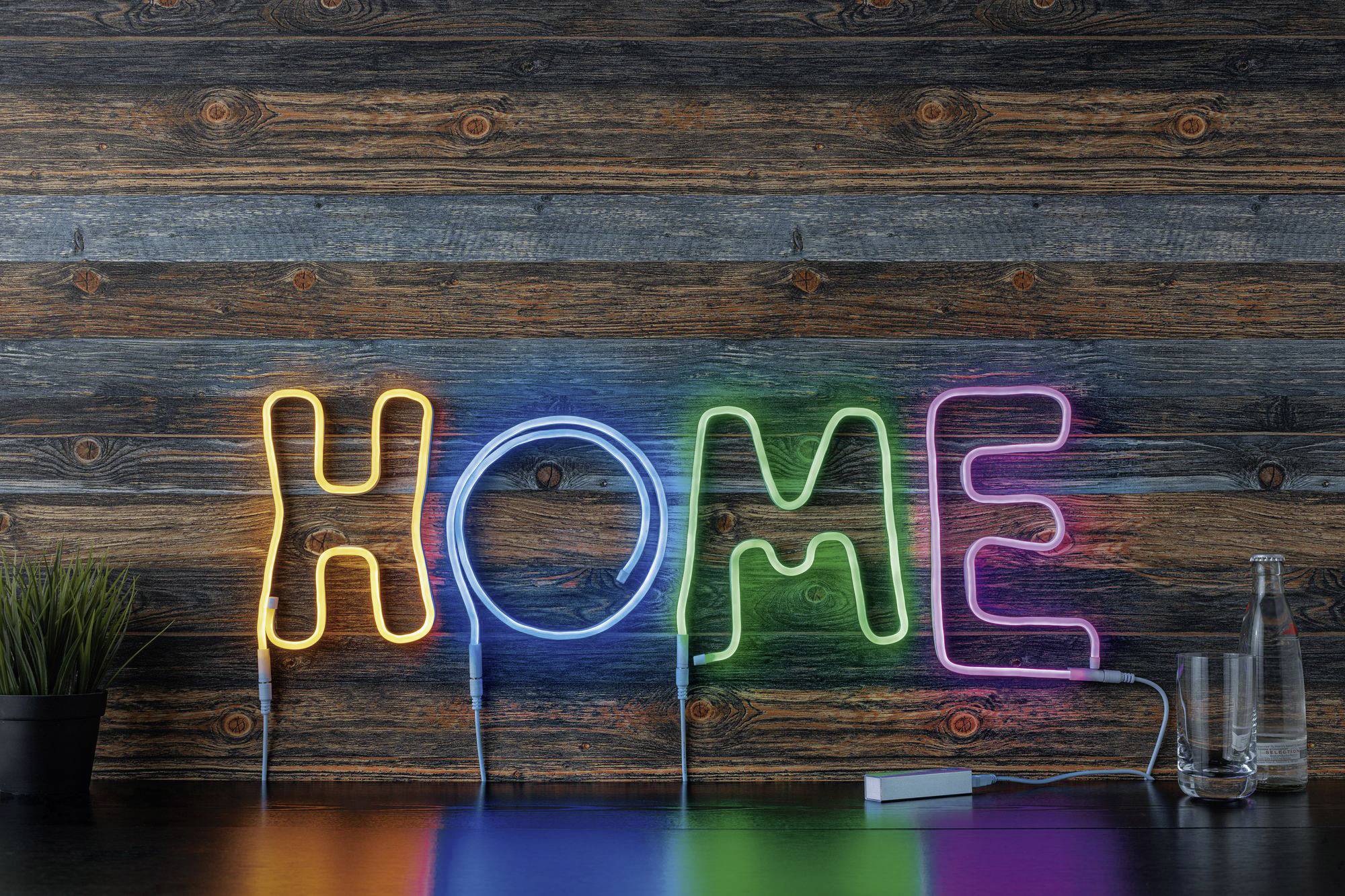 Insegna al neon con la parola 'HOME' in lettere colorate davanti a una parete di legno. A sinistra c'è una piccola pianta, a destra una bottiglia.