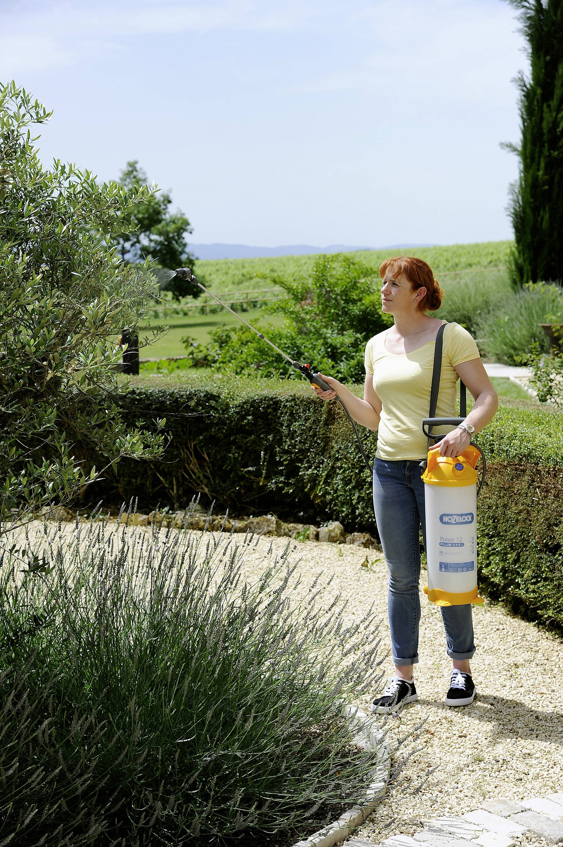 Donna con maglietta gialla che spruzza piante in un giardino con un contenitore per pesticidi. Sullo sfondo un ampio paesaggio.