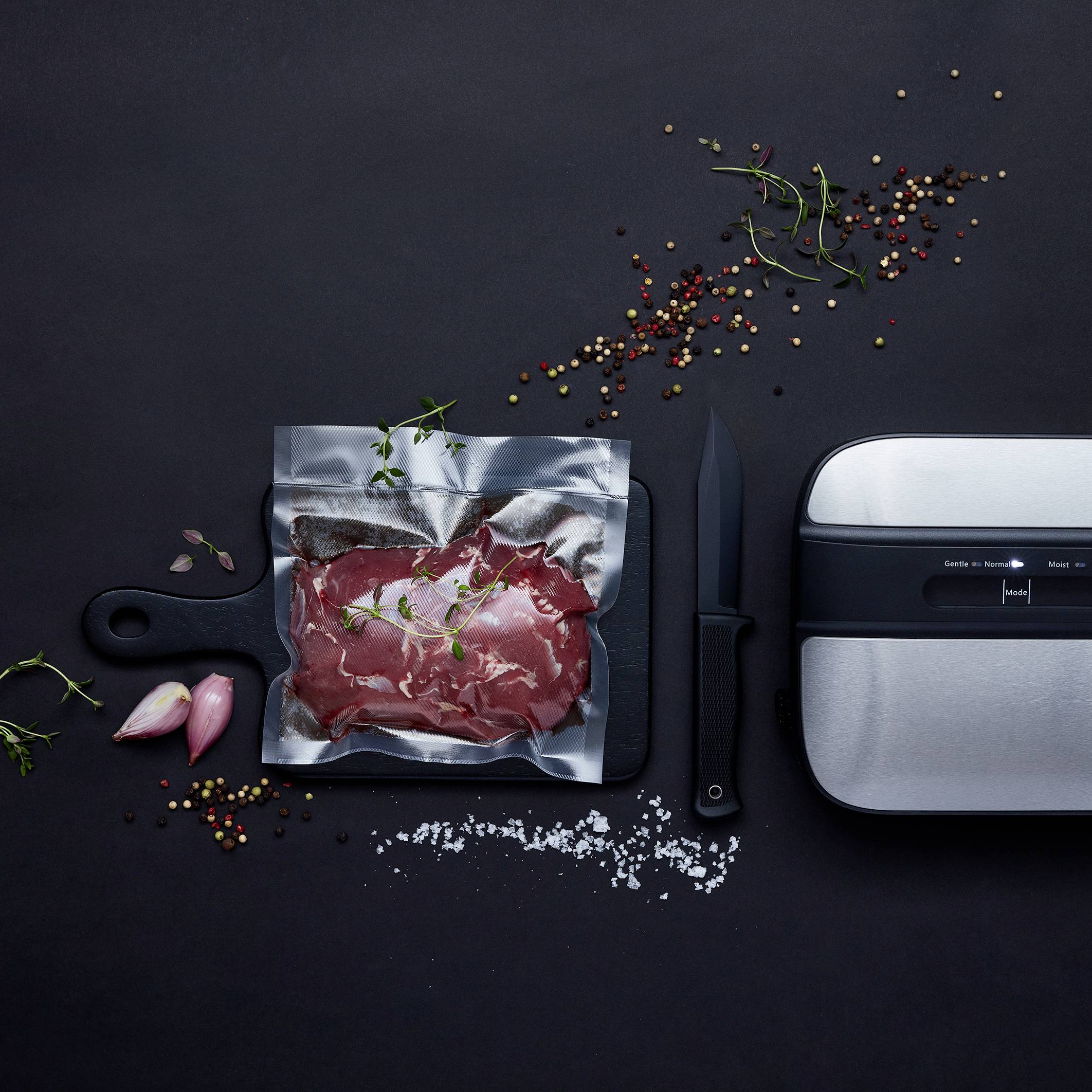 Un dispositivo per sottovuoto con un sacchetto pieno di carne su sfondo nero, circondato da grani di pepe, cipolla ed erbe aromatiche.