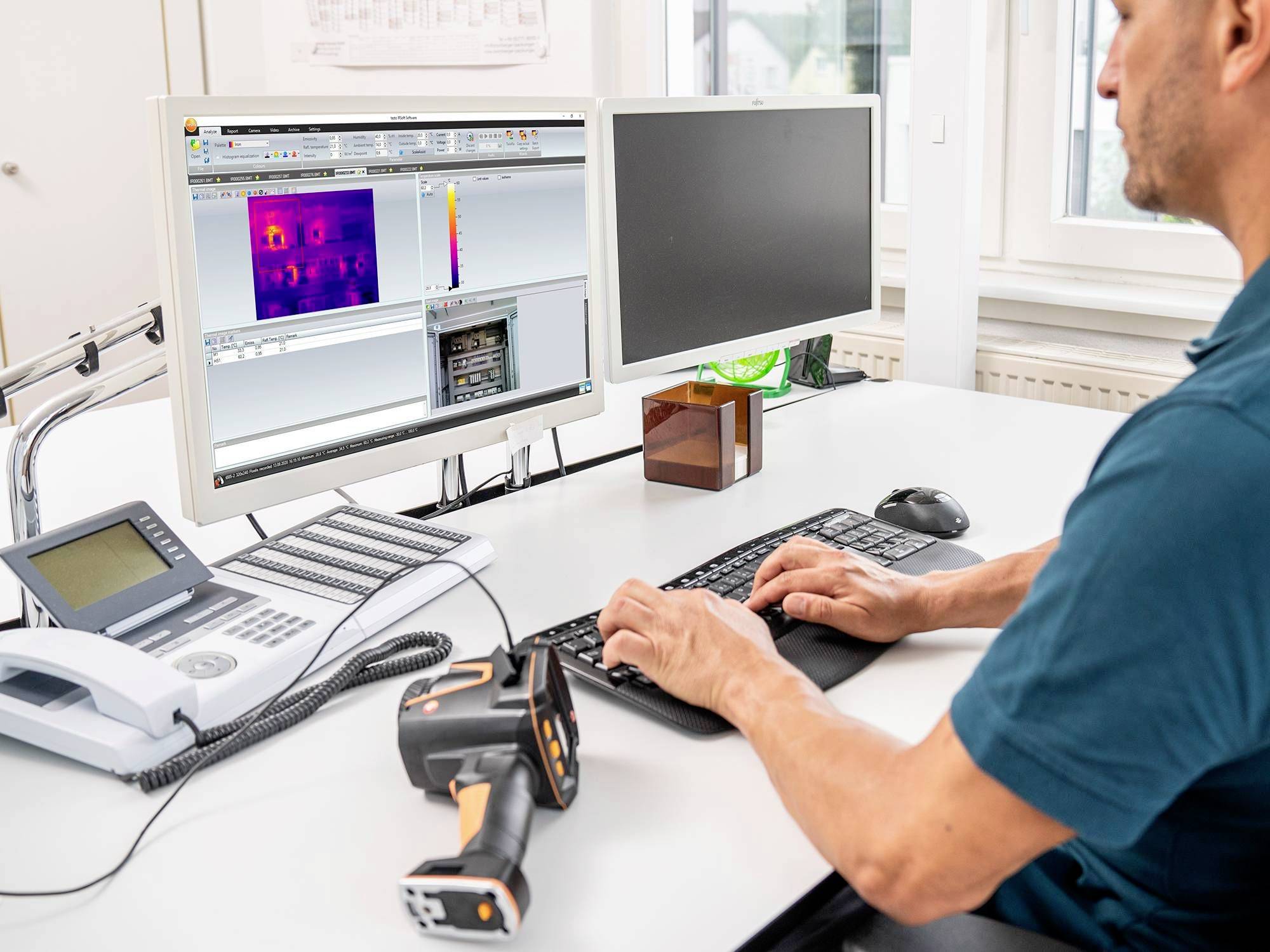 Un uomo è seduto a una scrivania e analizza dati termografici su uno schermo del computer. Accanto a lui è posizionata una fotocamera a infrarossi.