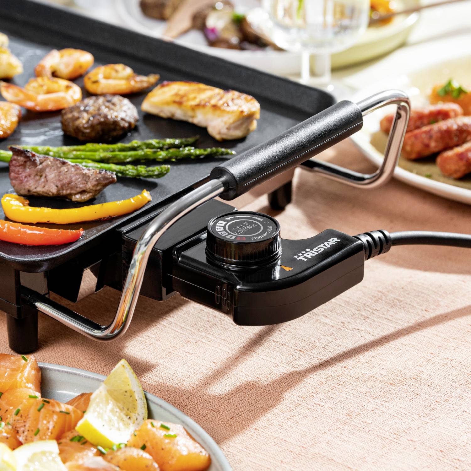Piastra elettrica per grigliare con verdure e carne grigliate su un tavolo con altri piatti. Il focus è sul cibo e la preparazione.