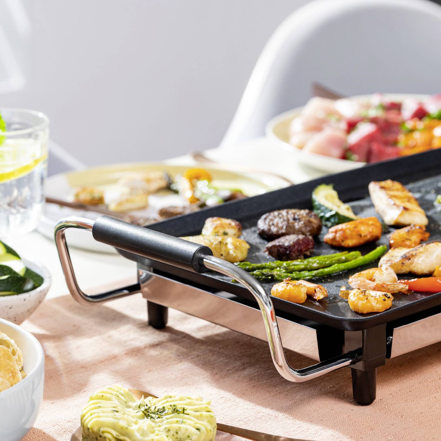Una piastra per grigliare con verdure grigliate, gamberi e pollo su un tavolo apparecchiato con insalata e bevanda al limone, nel complesso un pasto estivo.