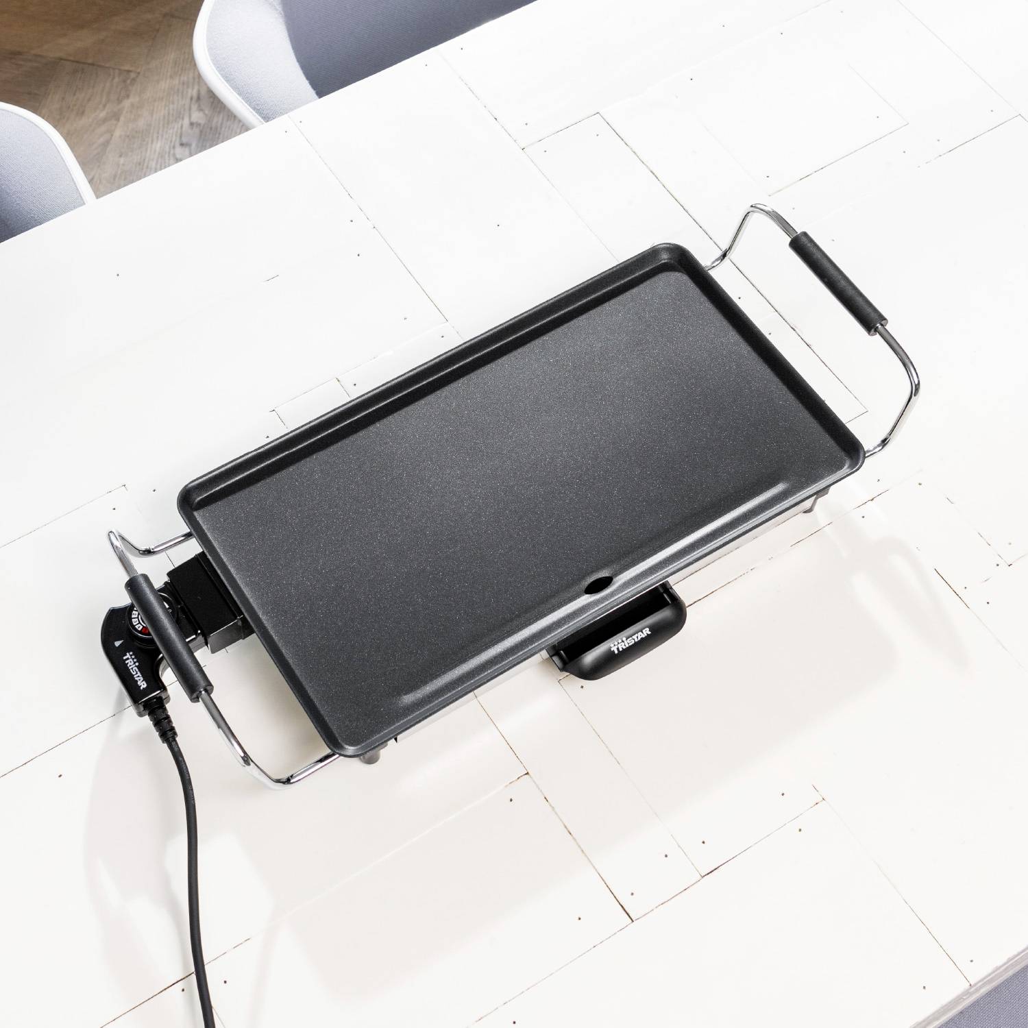 Un grill elettrico da tavolo con superficie antiaderente, posizionato su un tavolo bianco. Sullo sfondo sono visibili dei rivestimenti per sedie.