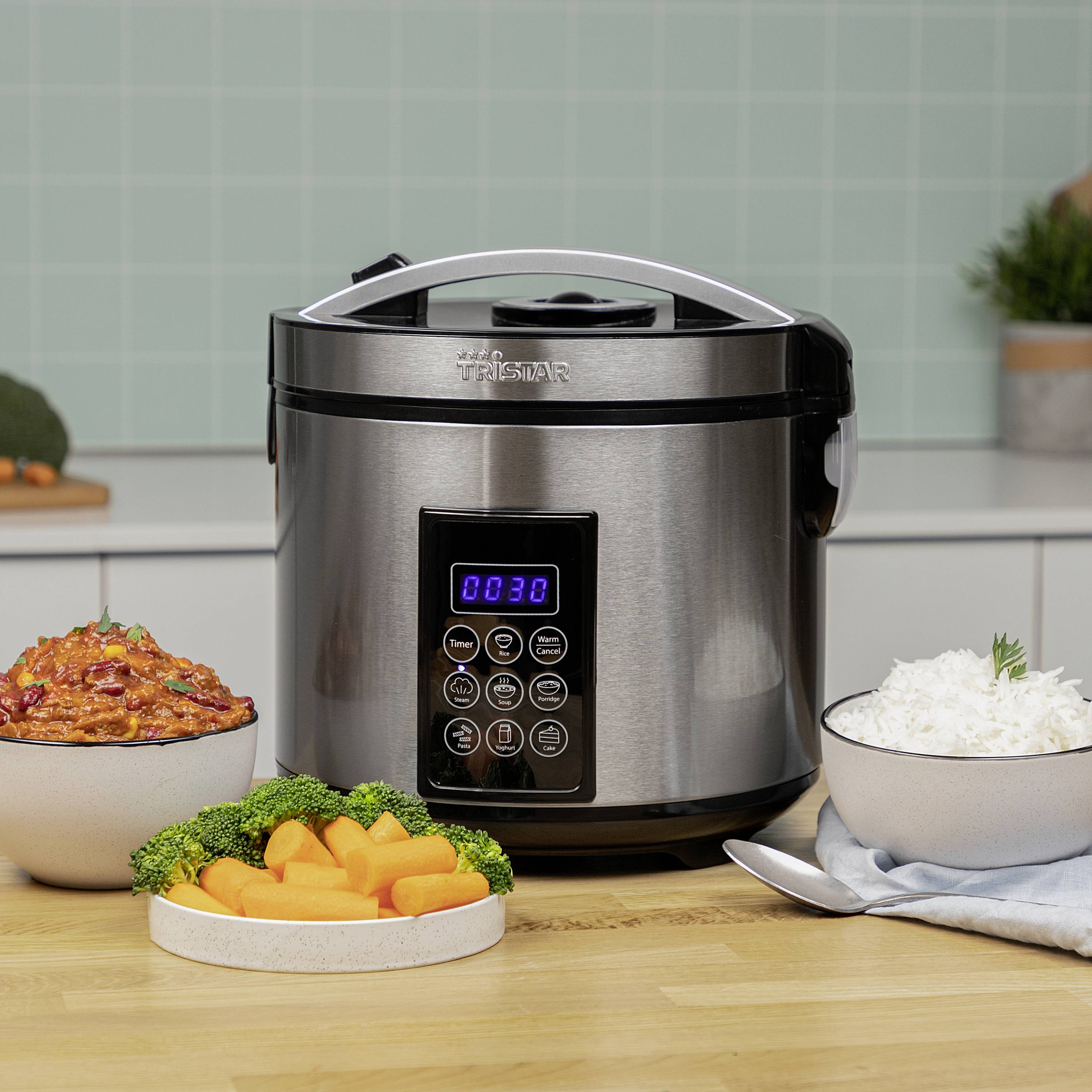 Multicooker con display digitale in una cucina. A sinistra curry di verdure, a destra una ciotola di riso. Sullo sfondo piastrelle blu.