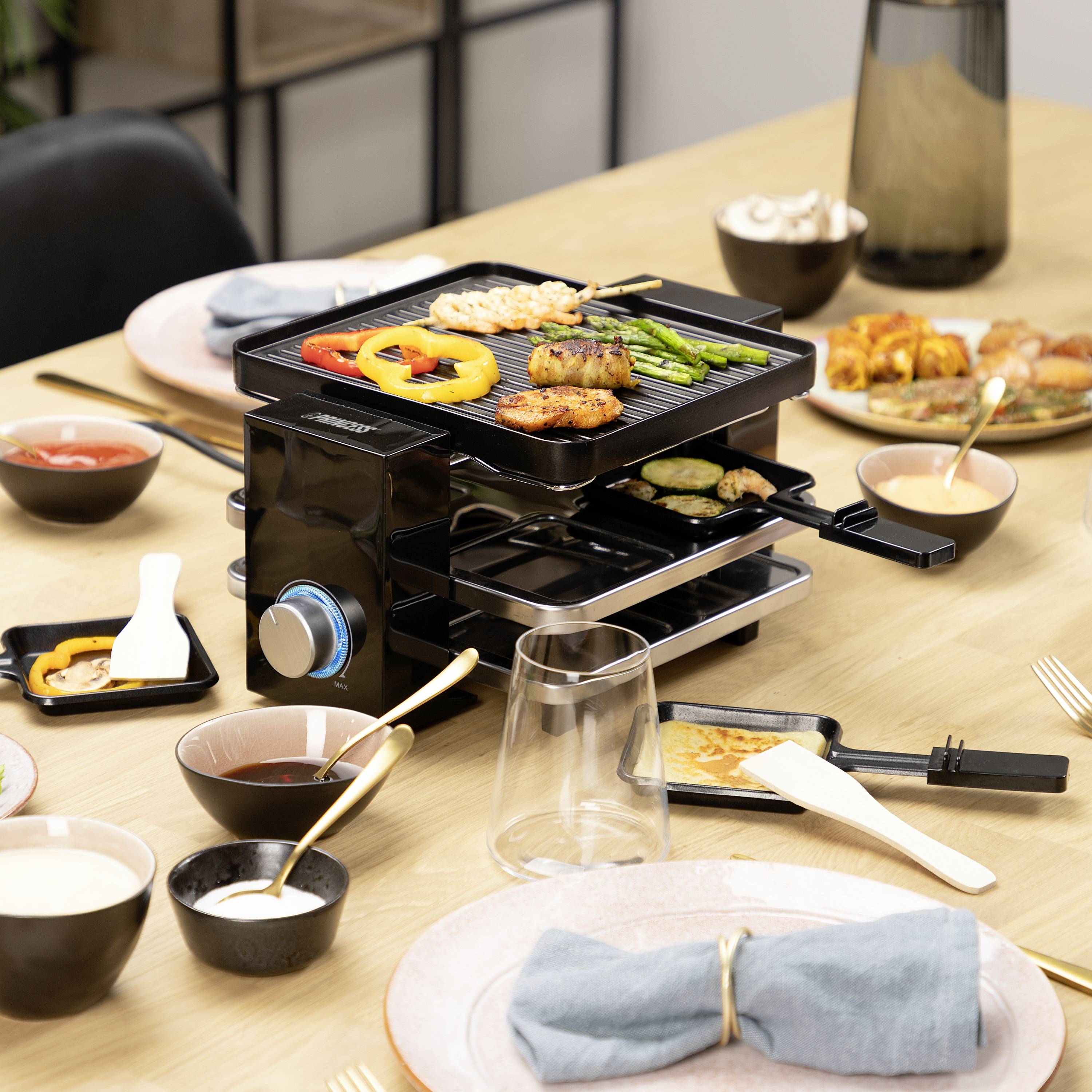 Un barbecue da tavolo con verdure e carne, circondato da un tavolo apparecchiato con piatti, ciotole e bevande in una sala da pranzo.