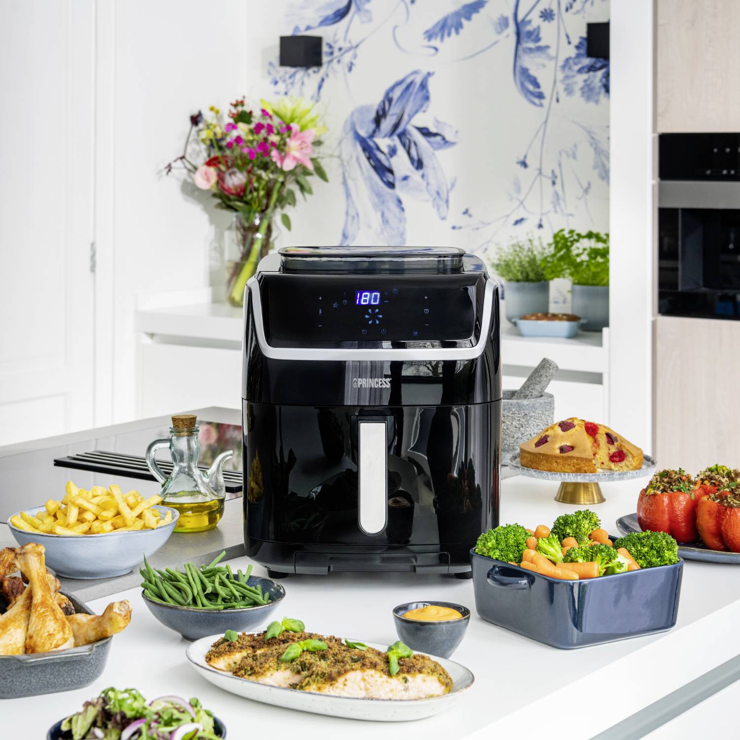 'Un'airfryer su un piano da lavoro della cucina, circondato da vari cibi come patatine fritte, pollo, peperoni ripieni e insalata.'