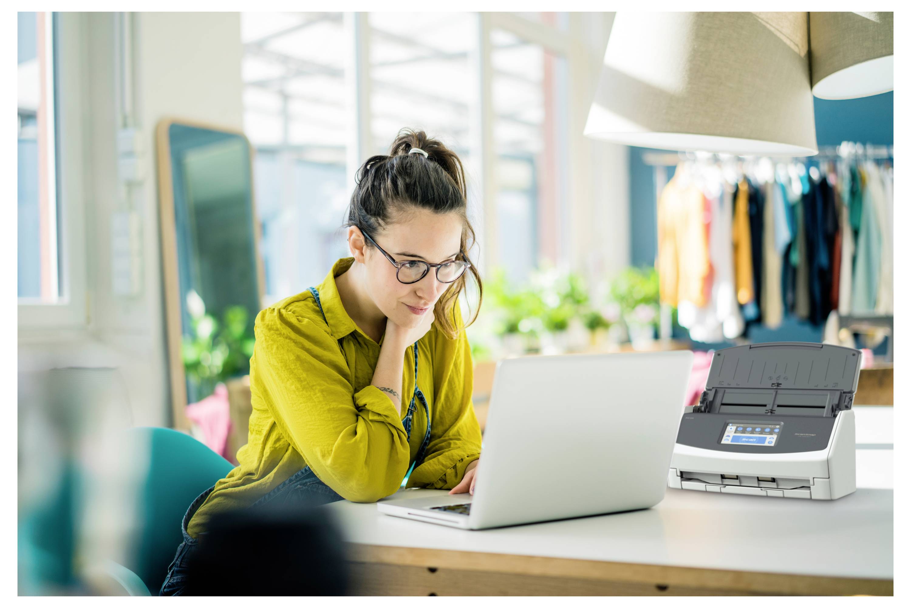 Una donna in un ufficio moderno e luminoso utilizza un laptop su una scrivania bianca. Una stampante è posizionata accanto a lei. Degli appendiabiti sono visibili sullo sfondo.