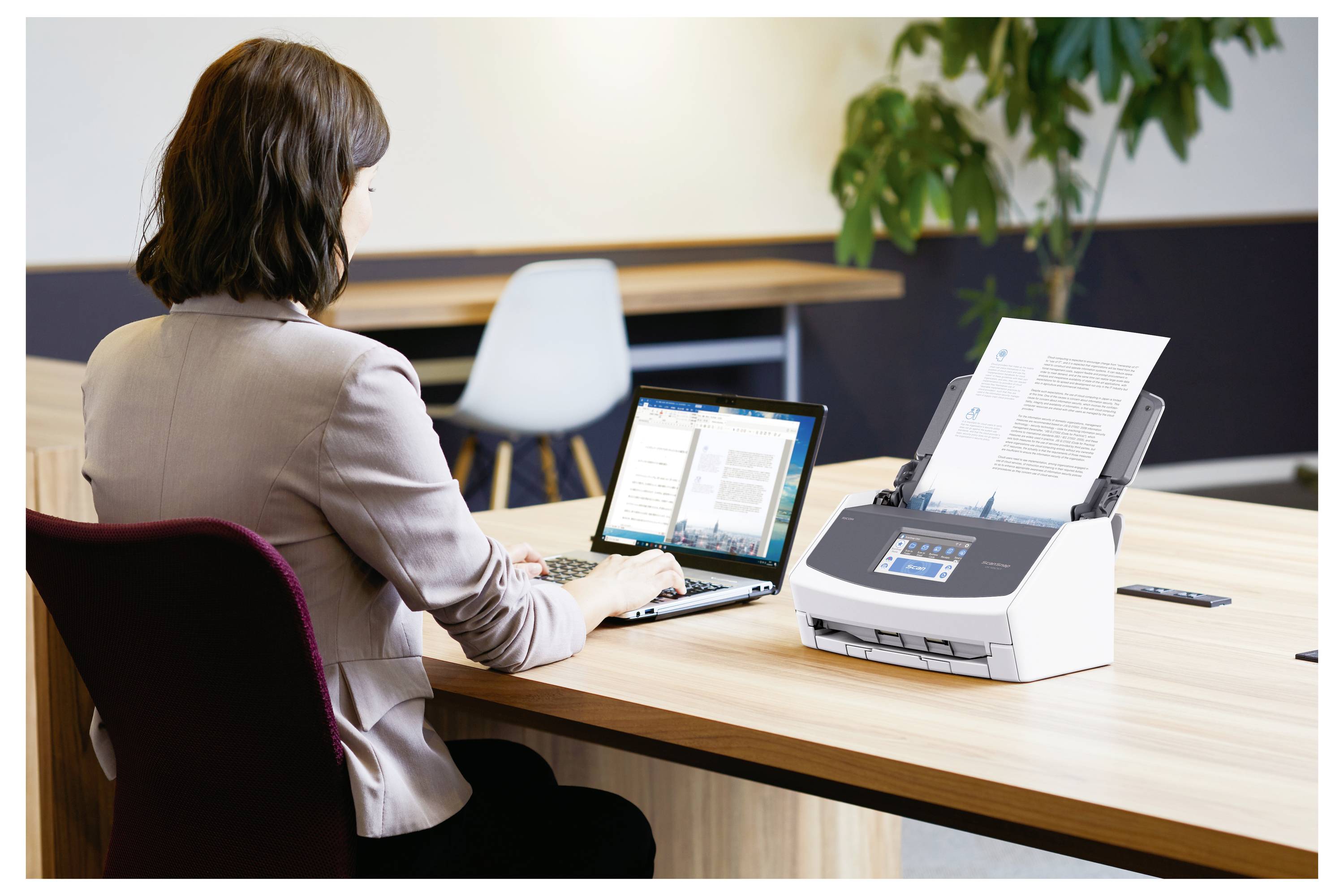 Una donna utilizza un laptop su una scrivania, con uno scanner a alimentazione automatica alla sua destra che sta acquisendo documenti. Ambiente di ufficio con un'altra scrivania.