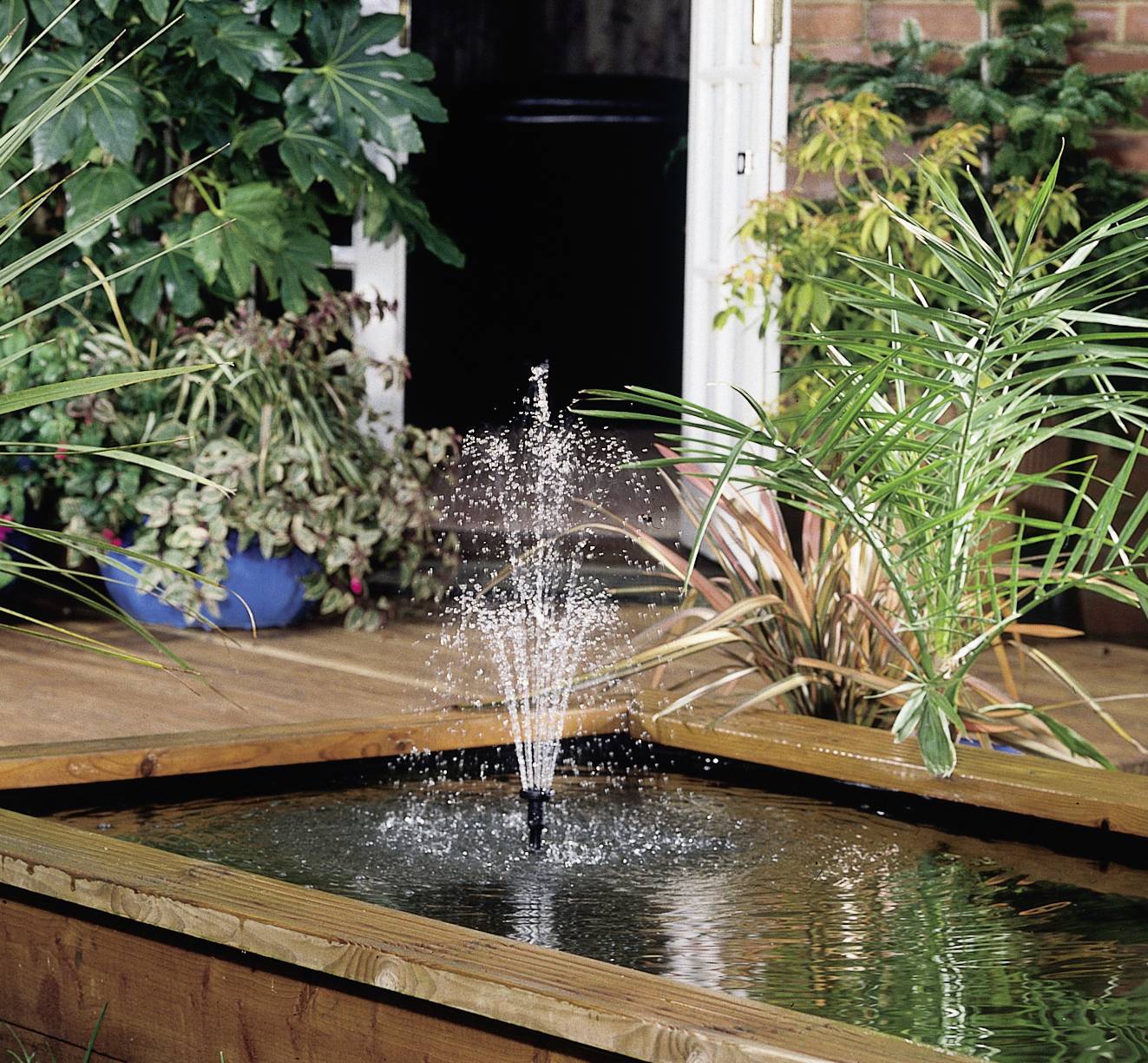 Una piccola fontana in un contenitore di legno rettangolare su una terrazza, circondato da grandi piante verdi in vasi.
