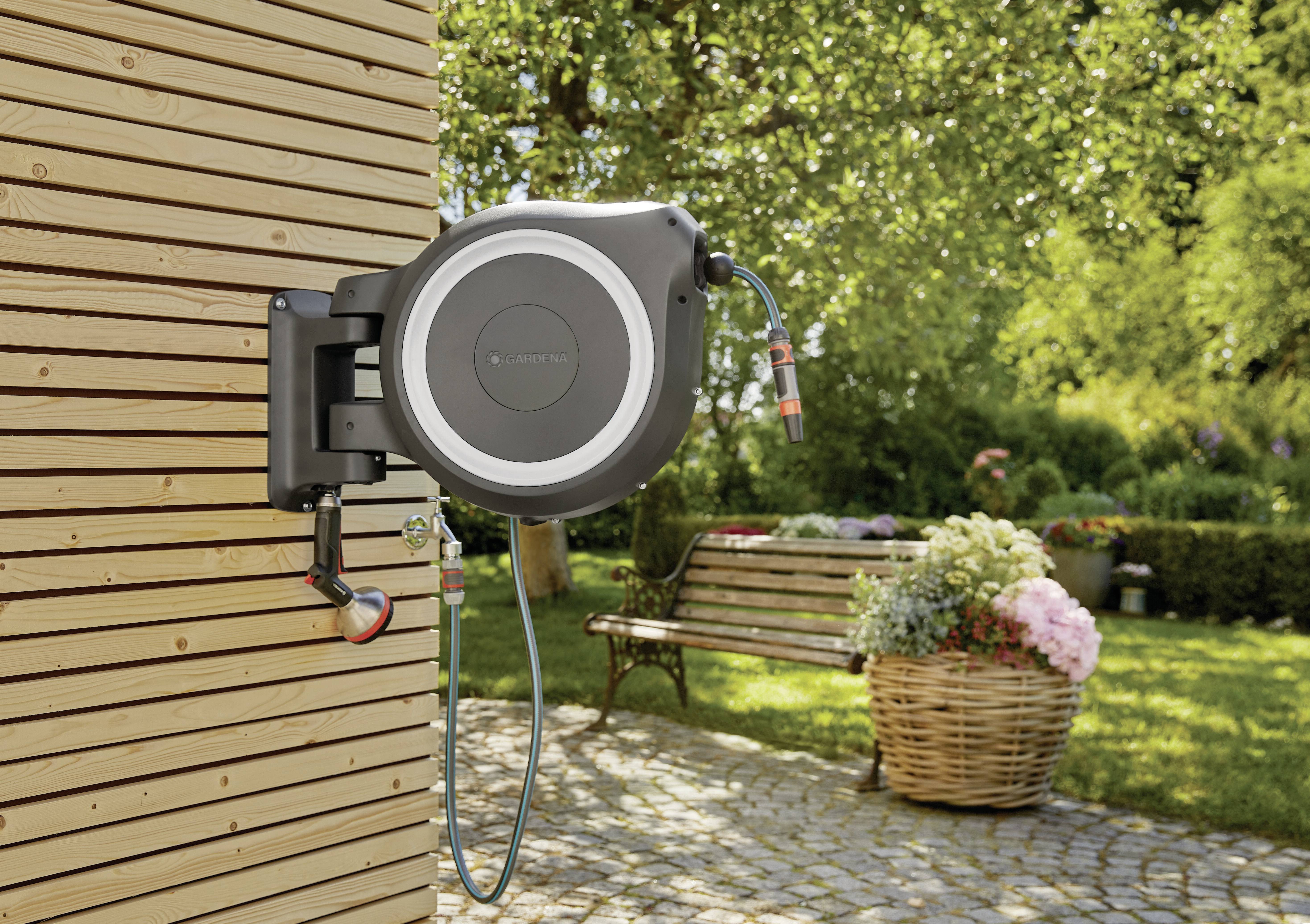 Un avvolgitubo automatico è montato su una parete in legno. Sullo sfondo si trova un giardino con una panchina e dei fiori.