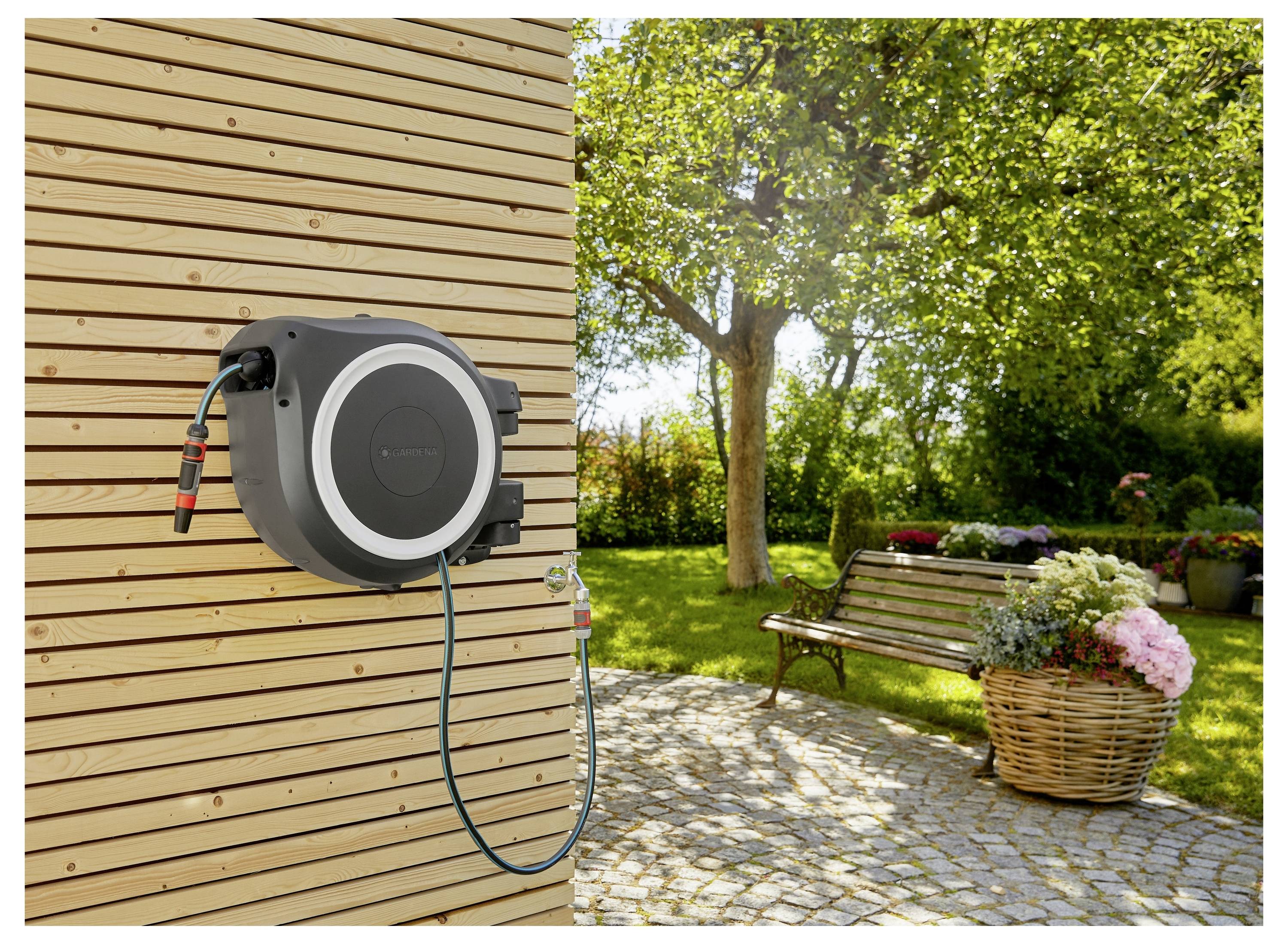 Un avvolgitubo da giardino montato su una parete in legno con un patio pavimentato, circondato da vegetazione, alberi, una panchina in legno e un cesto di fiori.