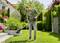Un uomo in jeans e una camicia verde sta tagliando l'erba in un giardino ben curato. Sullo sfondo si vedono cespugli e fiori.