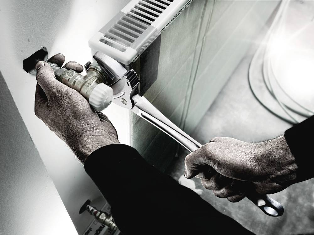 Una persona sta stringendo una valvola su un radiatore con una chiave inglese. La luce diurna entra nella stanza da destra.