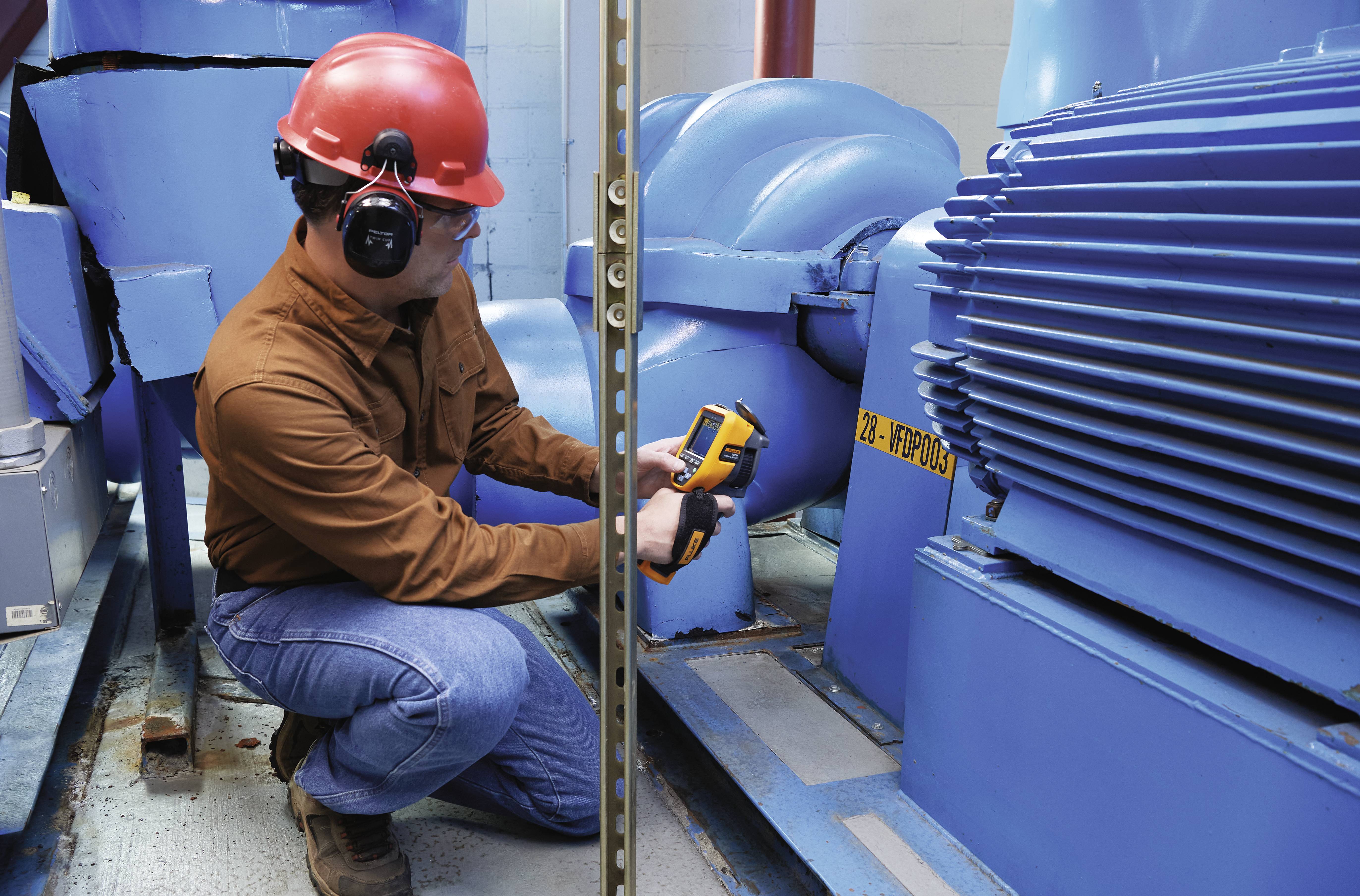 Un tecnico con casco protettivo e protezioni per l'udito misura la temperatura di un grande motore blu utilizzando un termometro a infrarossi.