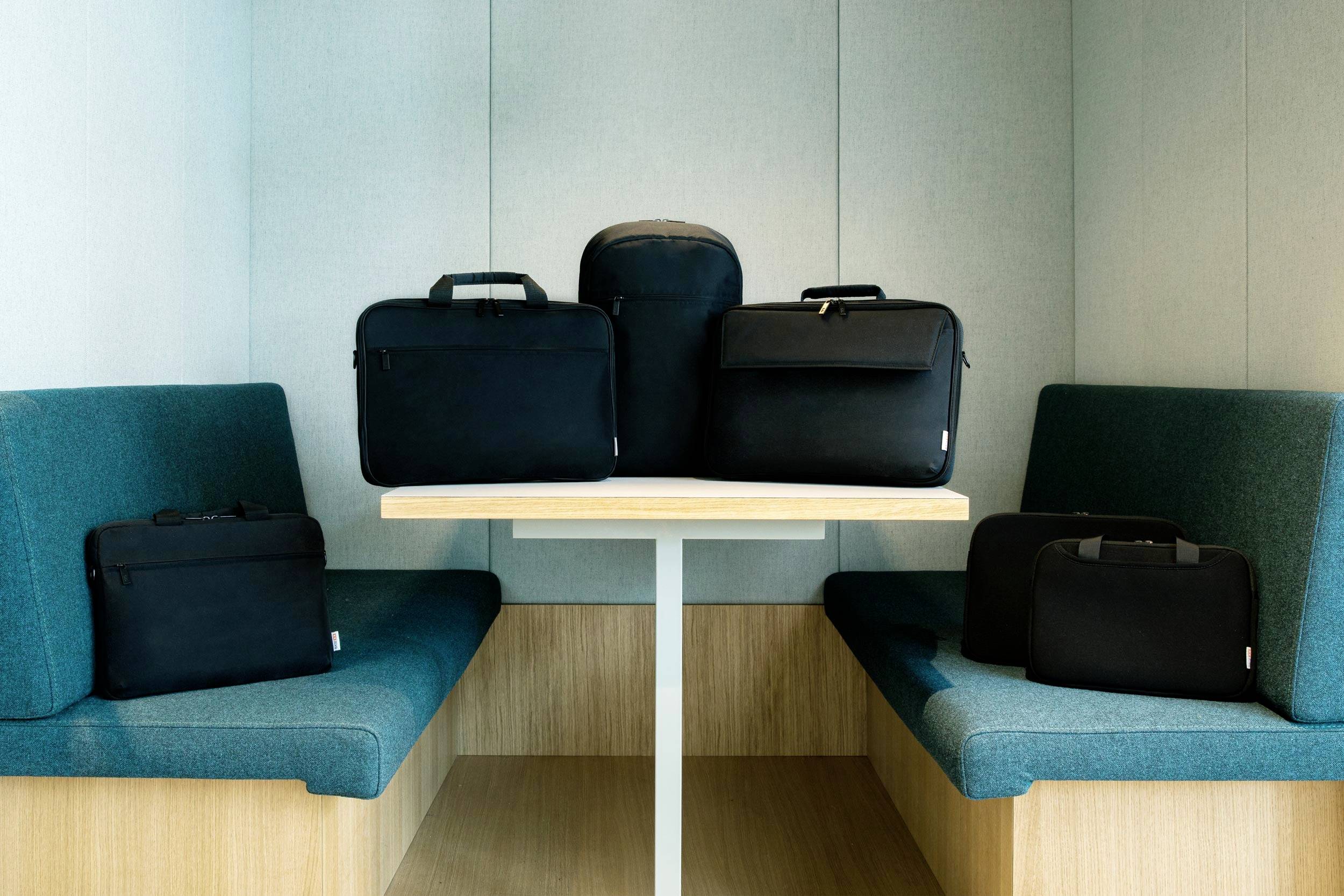 Borse per laptop di varie dimensioni su un tavolo tra due zone sedute. Ambiente moderno e minimalista.