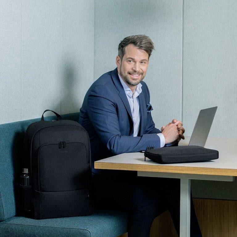 Uomo in giacca e cravatta sorride seduto a un tavolo con un laptop; zaino e bottiglia d'acqua accanto. La scena suggerisce un colloquio di lavoro o una situazione professionale.