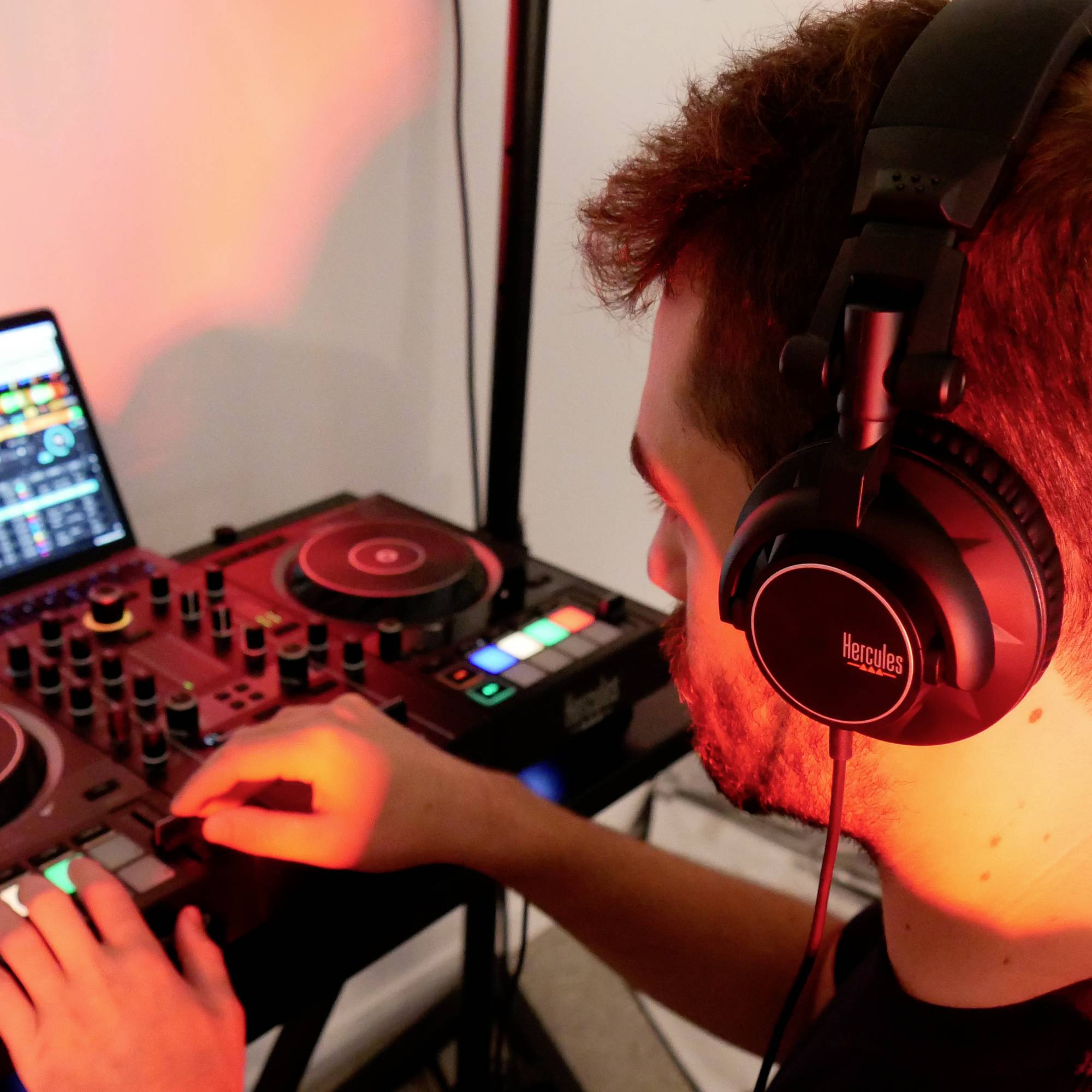 Un DJ mixa musica su un controller per DJ, con le cuffie sulle orecchie, concentrato nell'adattare i controlli; sullo sfondo uno schermo.