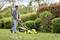 Un uomo spinge un tosaerba giallo su un prato verde. Cespugli e alberi sono sullo sfondo.