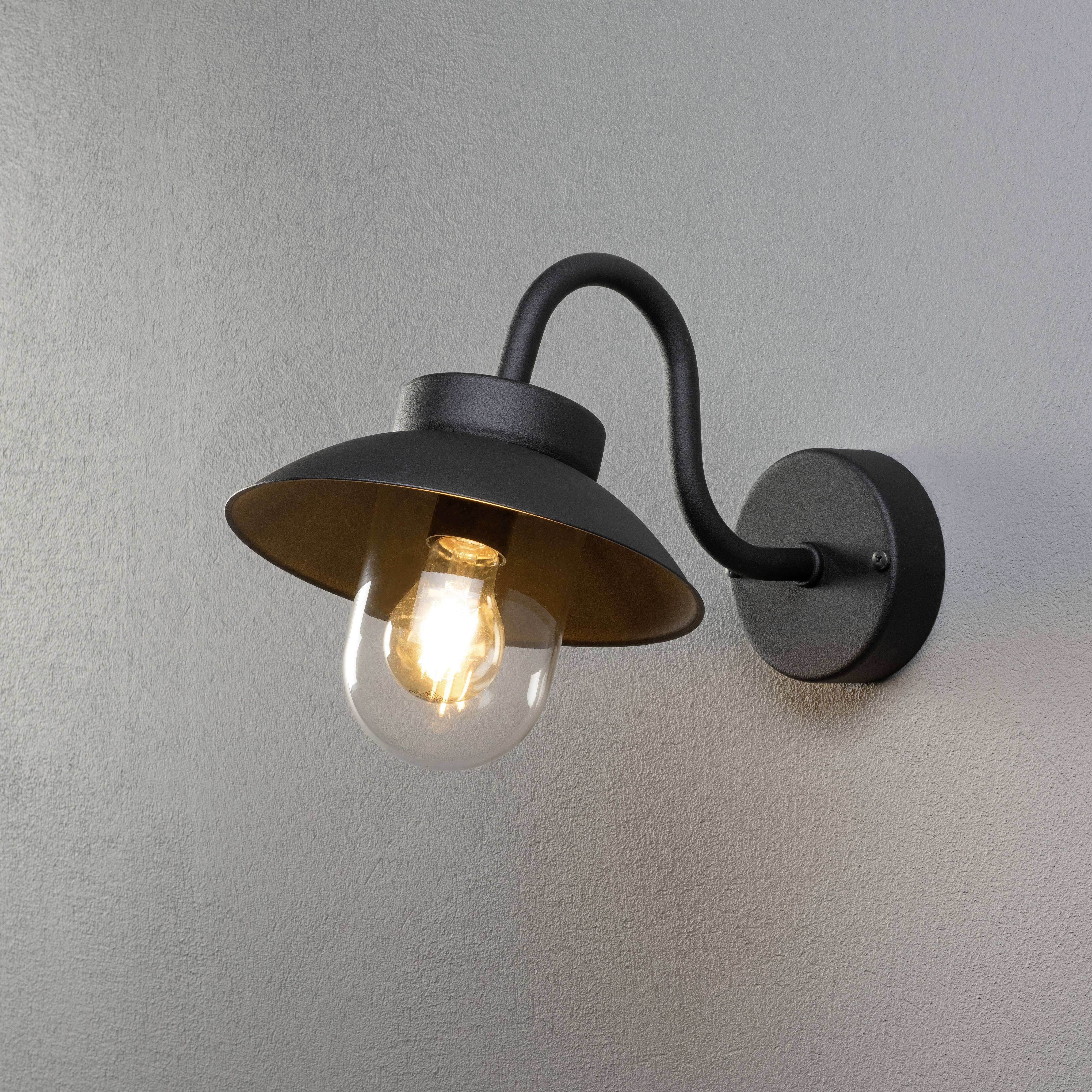 Applique da parete nera con braccio curvo e lampadina a vista, fissata a una parete grigia e che emana una luce calda.