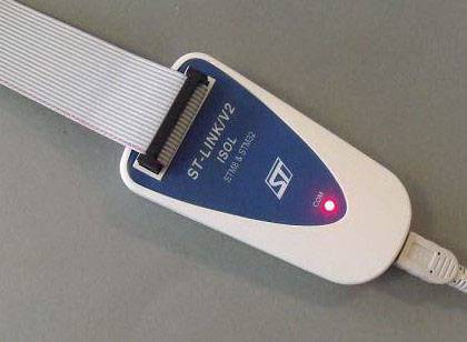 Un debugger ST-LINK/V2-ISOL con etichetta blu e luce LED rossa su un tavolo. Collegato con un cavo piatto bianco e un cavo USB.