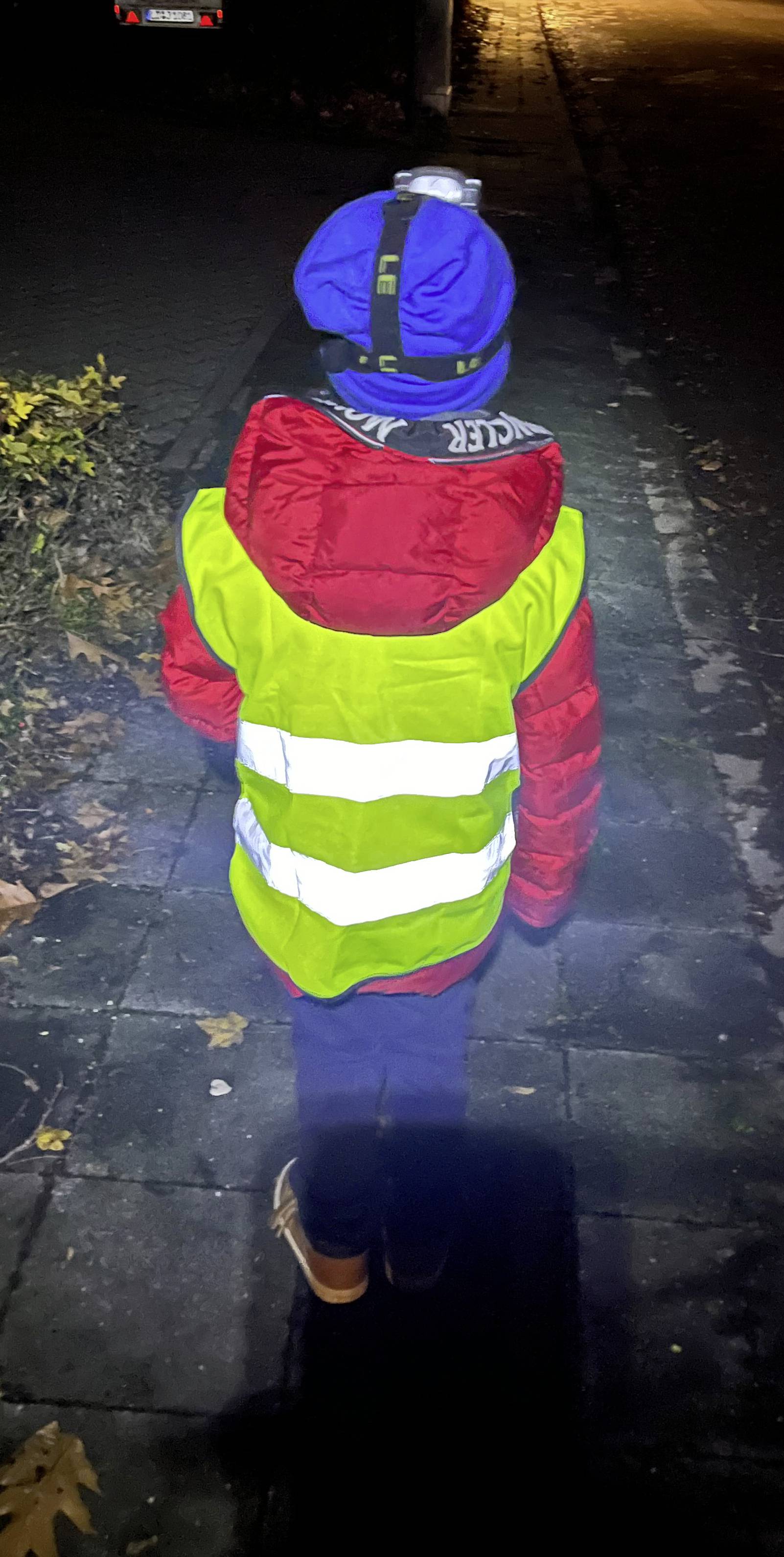 Un bambino con un berretto blu e una giacca rossa indossa un giubbotto catarifrangente giallo e cammina su un marciapiede di notte.