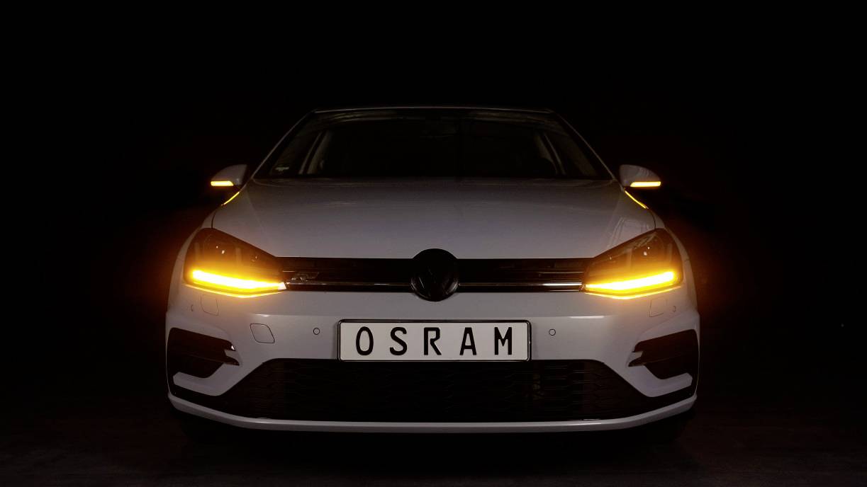 Auto bianco con fari distintivi color giallo brillante e frecce retrovisori sullo sfondo nero, targa 'OSRAM'.