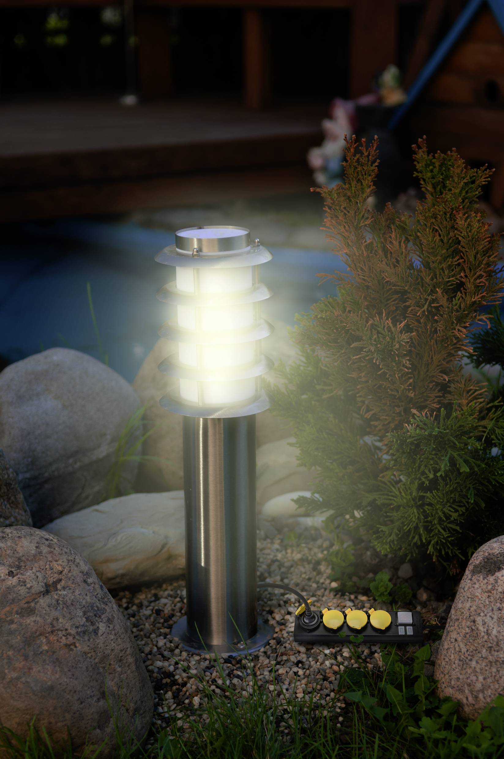 Lampada da giardino in alluminio illuminata su un'aiuola sassosa. Nelle vicinanze è presente un multipresa. Sullo sfondo sfocato.