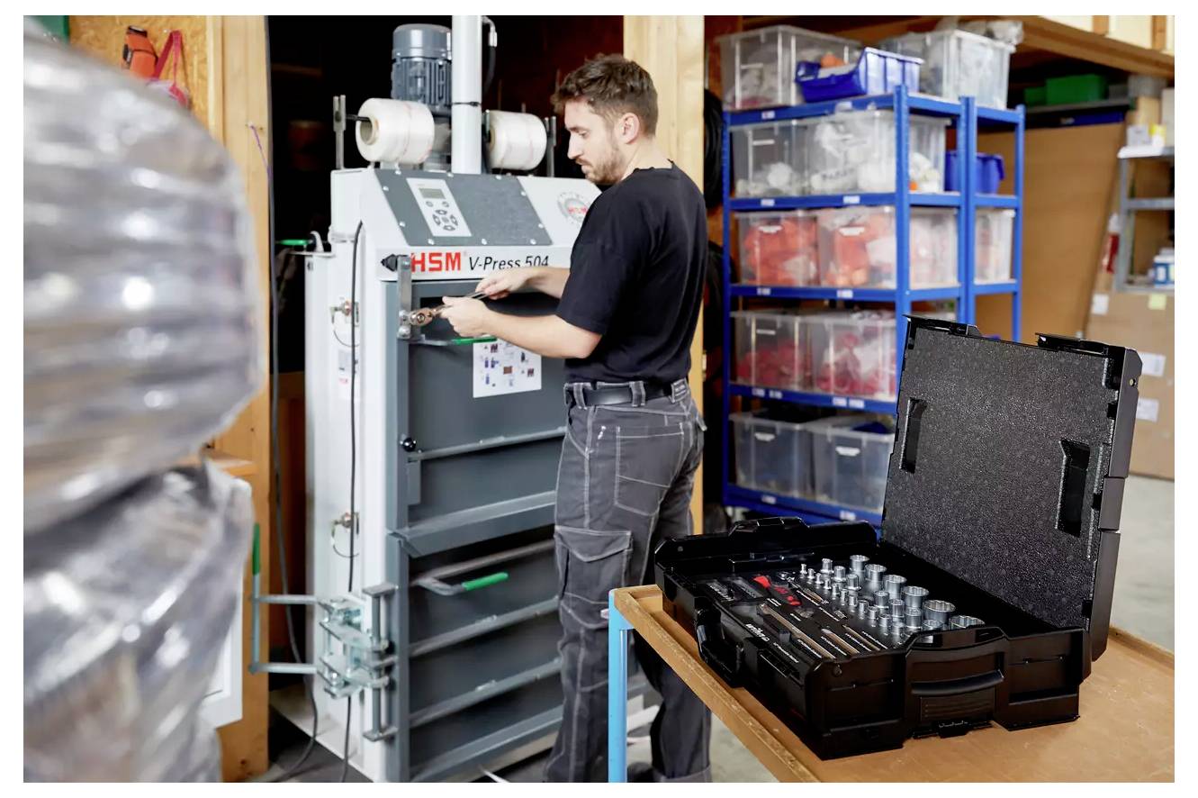 Una persona aziona una macchina per la pressione HSM in un'officina, con scaffali organizzati e un cassone degli attrezzi vicino, indicando un'attività lavorativa concentrata.