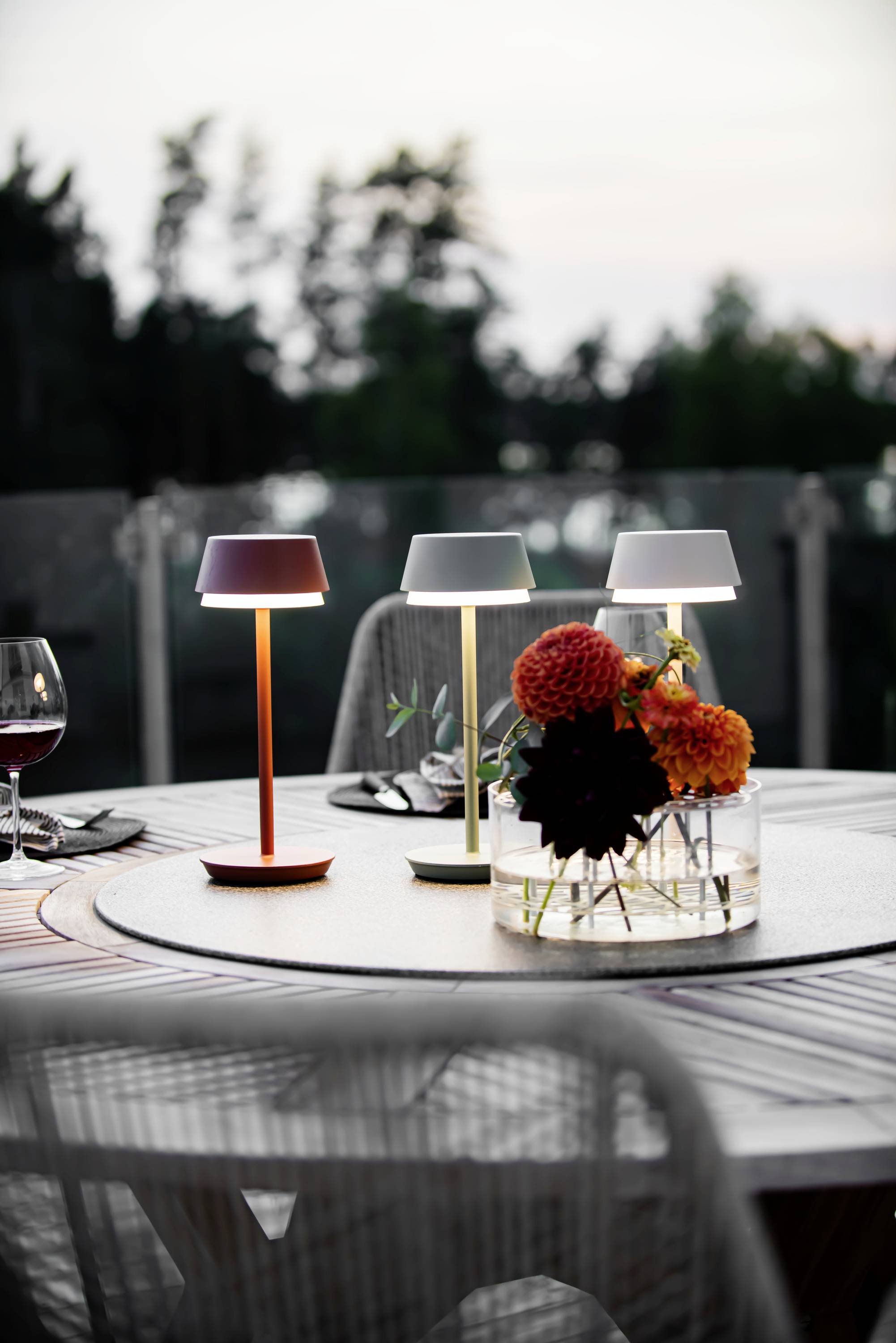 Un tavolo rotondo su una terrazza al tramonto, apparecchiato con bicchieri di vino, fiori colorati in un vaso e tre lampade da tavolo illuminate.