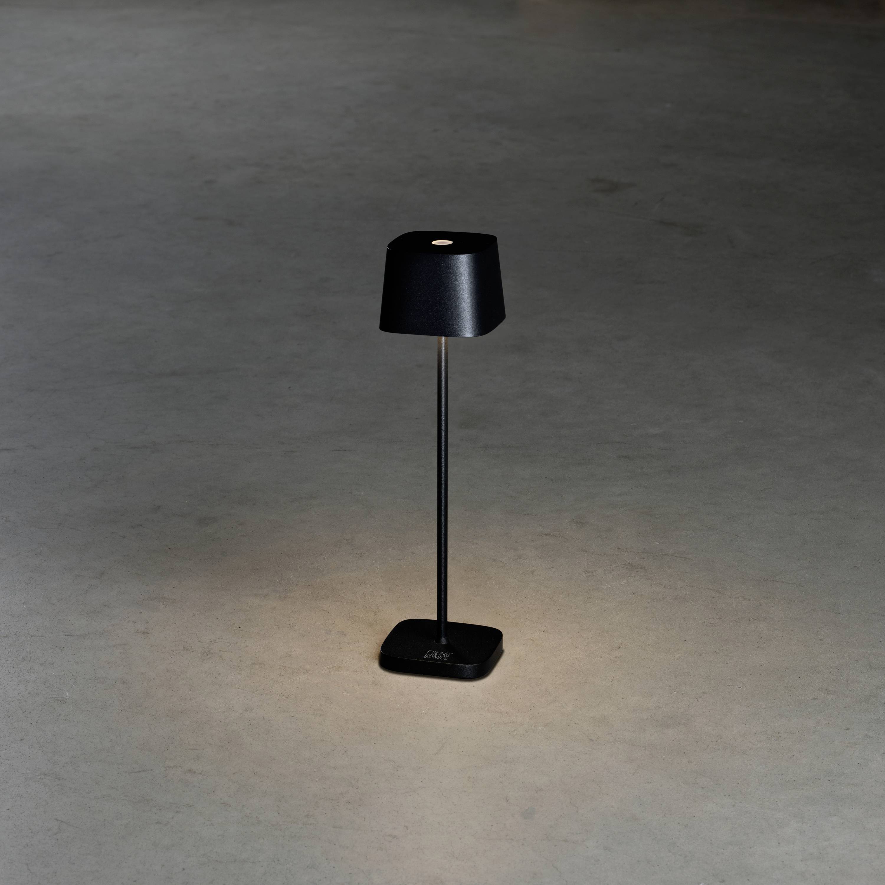 Lampada da tavolo nera dal design essenziale che illumina delicatamente il pavimento grigio circostante. Il paralume è a forma di cilindro.
