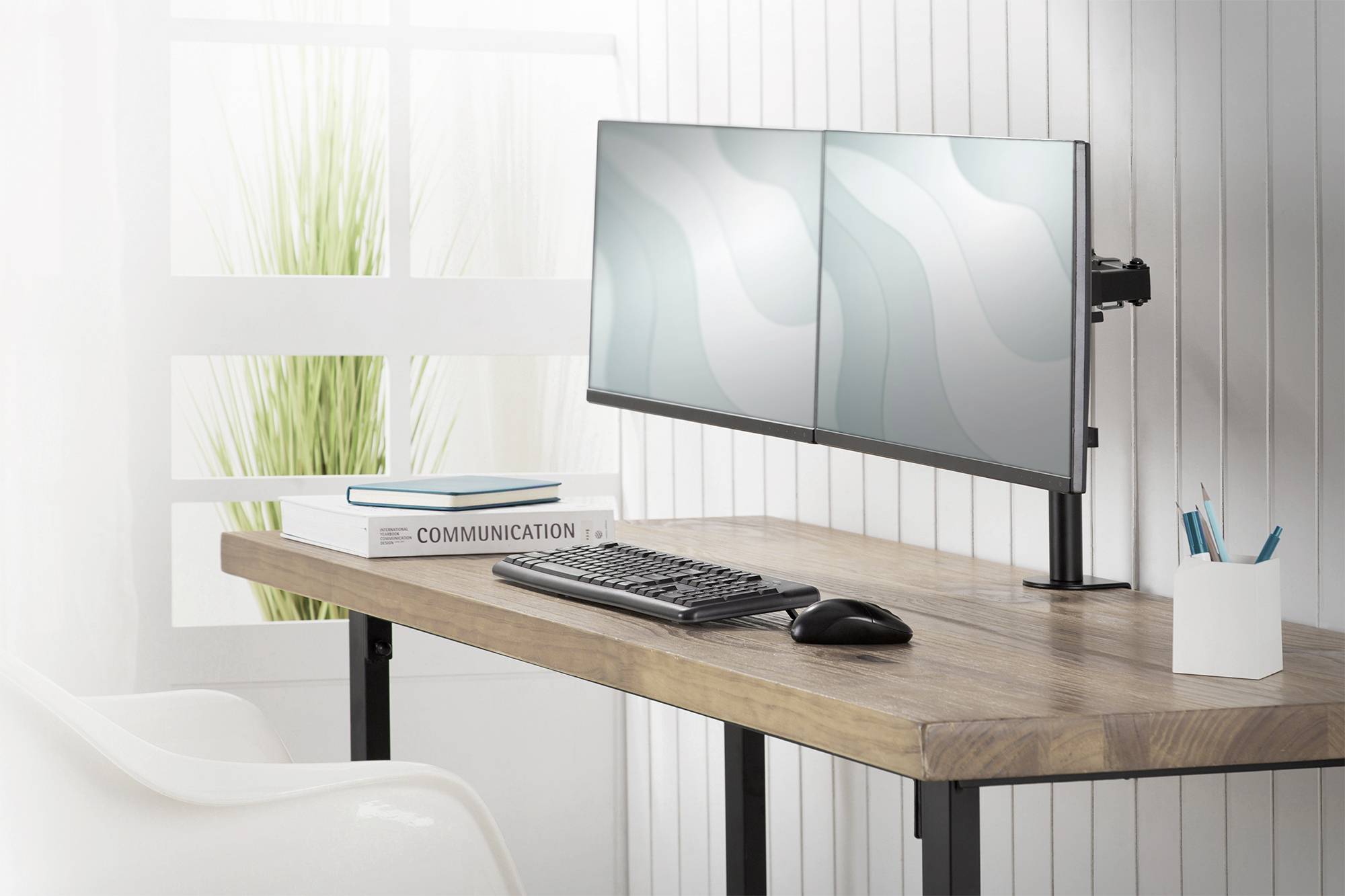 Una scrivania con un setup a doppio monitor, accanto una tastiera e un mouse. Un libro dal titolo 'Communication' è posizionato a sinistra.