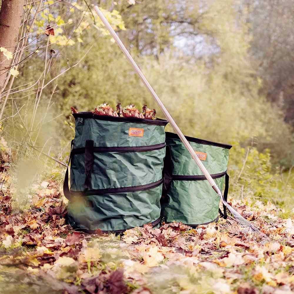 Due sacchi verdi per rifiuti da giardino, riempiti di foglie, sono posizionati su fogliame autunnale accanto a un albero; un rastrello è appoggiato loro vicino.