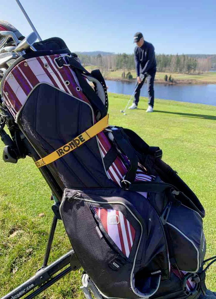 Borsa da golf in primo piano, golfista sullo sfondo davanti a uno stagno, pronto a colpire su un campo da golf soleggiato.