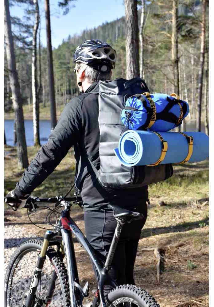 Una persona in bicicletta trasporta uno zaino grande con materassini blu e tende. Sta percorrendo un sentiero nel bosco che costeggia un lago.