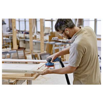 Un uomo che indossa protezioni auricolari utilizza una levigatrice elettrica su una cornice di legno in un laboratorio pieno di attrezzi e materiali per la lavorazione del legno.