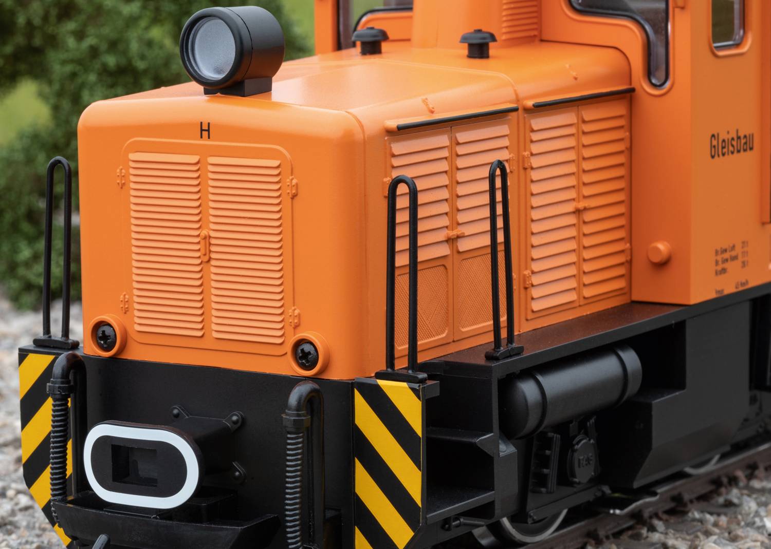 Locomotiva arancione con strisce nere, scritta 'Gleisbau', parte anteriore dettagliata, ambiente circostante con vegetazione verde visibile.