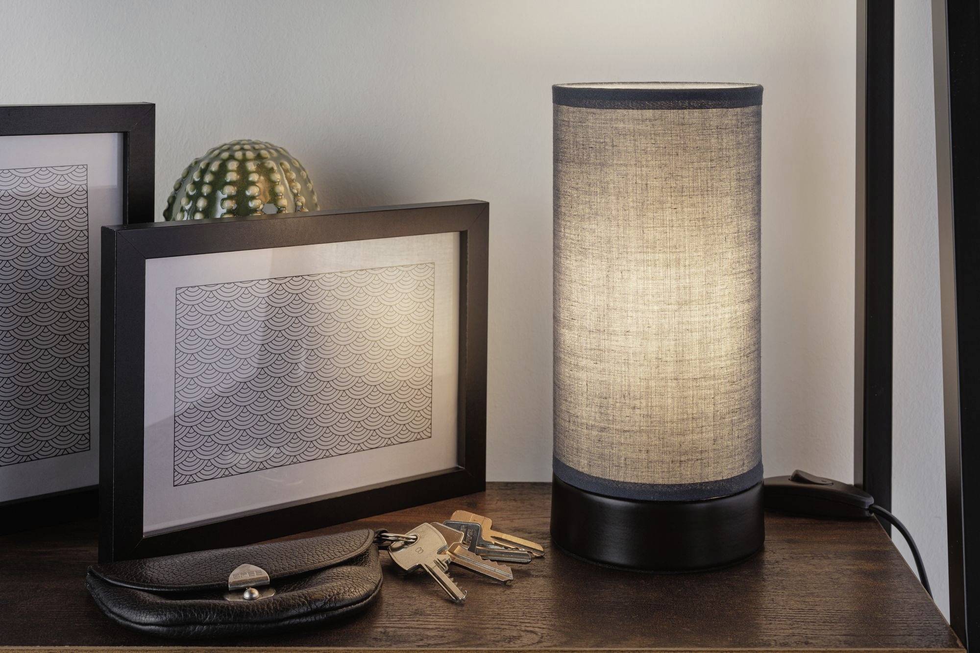 Una lampada da tavolo illumina accanto a due cornici con immagini e un cactus. Davanti, su un tavolo, è posato un portachiavi con delle chiavi.