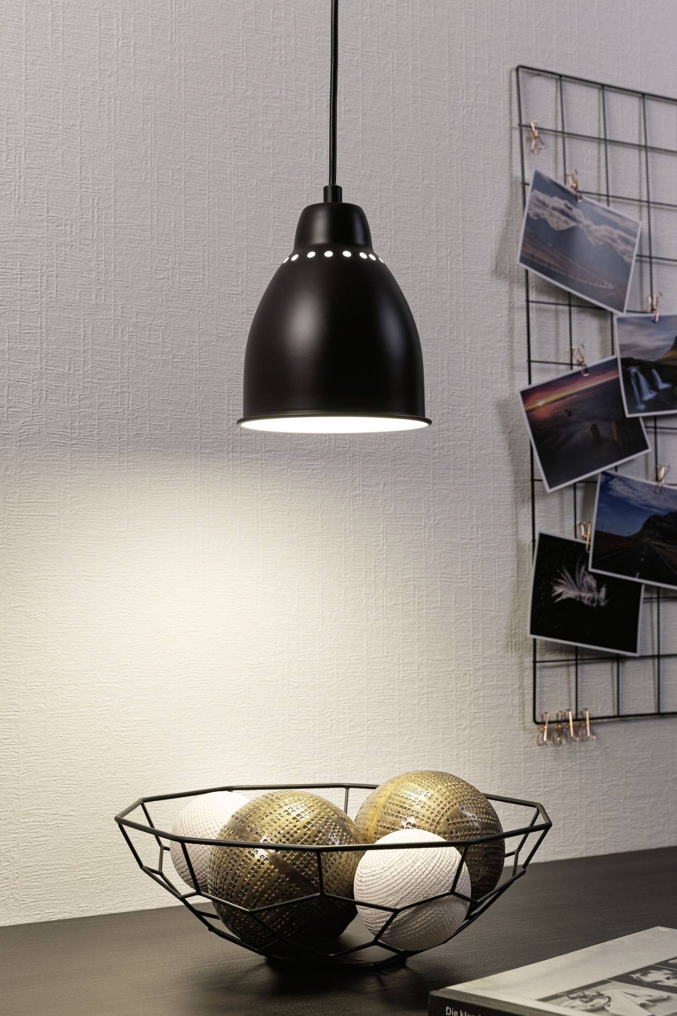 Lampada a sospensione nera illumina un tavolo con un cestino di fili metallici che contiene sfere decorative oro e bianche. Sullo sfondo sono appese delle fotografie.