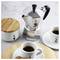 Un caffettiera moka accanto a due tazzine di espresso su piattini, accompagnata da un piccolo vasetto con un coperchio in legno, il tutto su una superficie chiara.