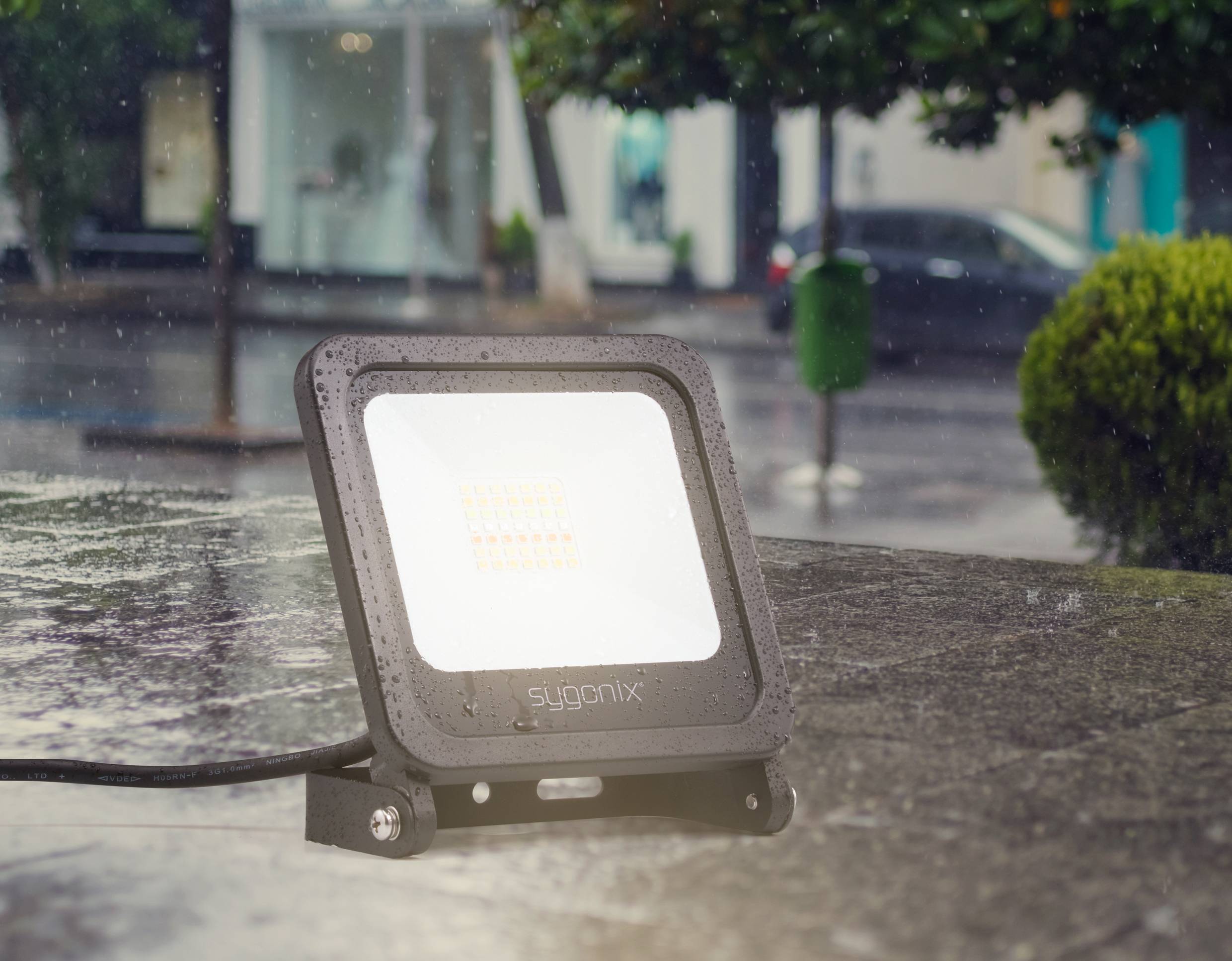 Una lampada LED da inondazione nera è posizionata su un tavolo all'aperto sotto la pioggia. Sullo sfondo si vedono degli alberi e una strada bagnata dalla pioggia.