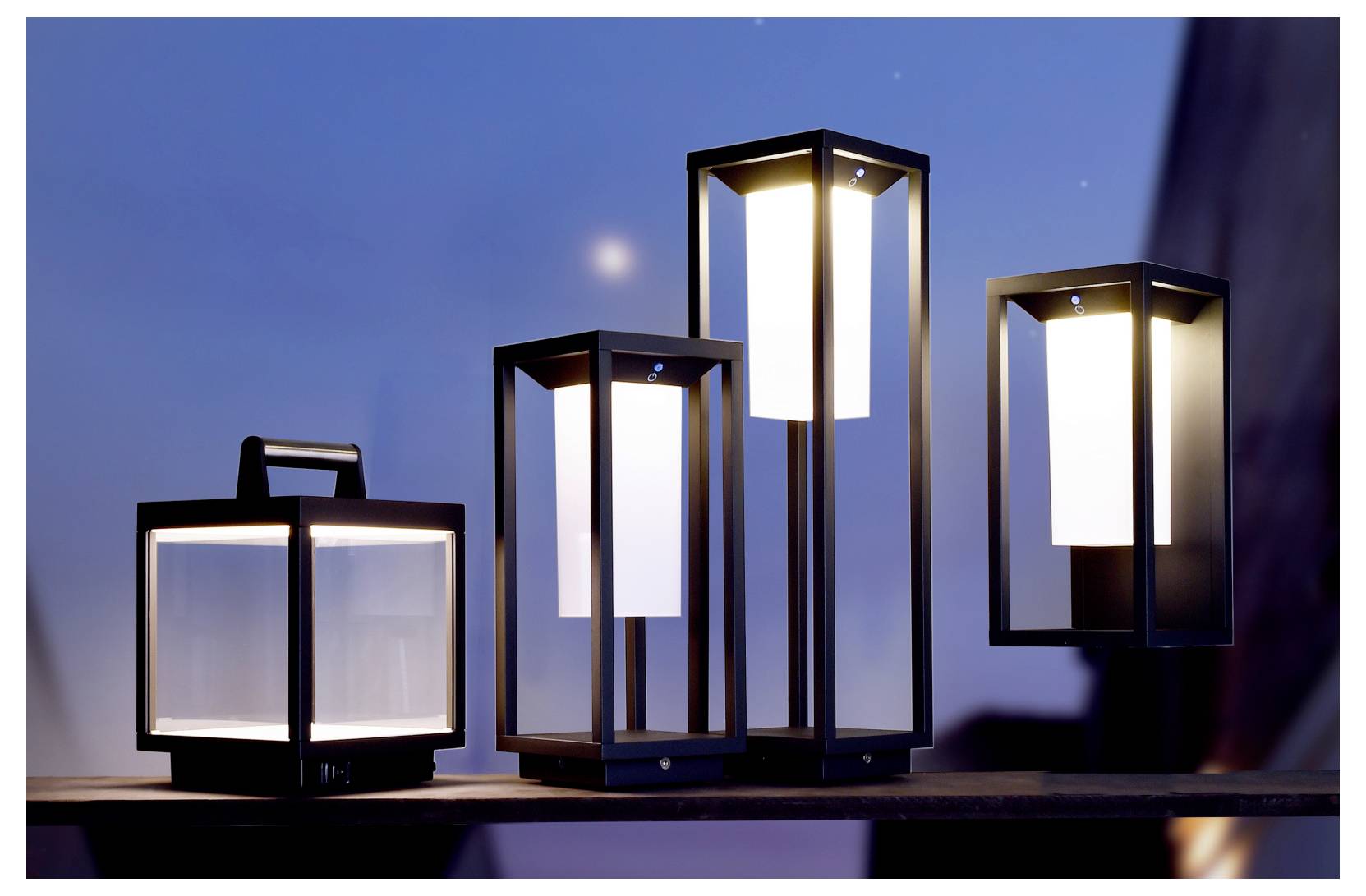 Quattro lanterne da esterno accese su una mensola contro un cielo crepuscolare, con telai neri moderni e pannelli bianchi luminosi.