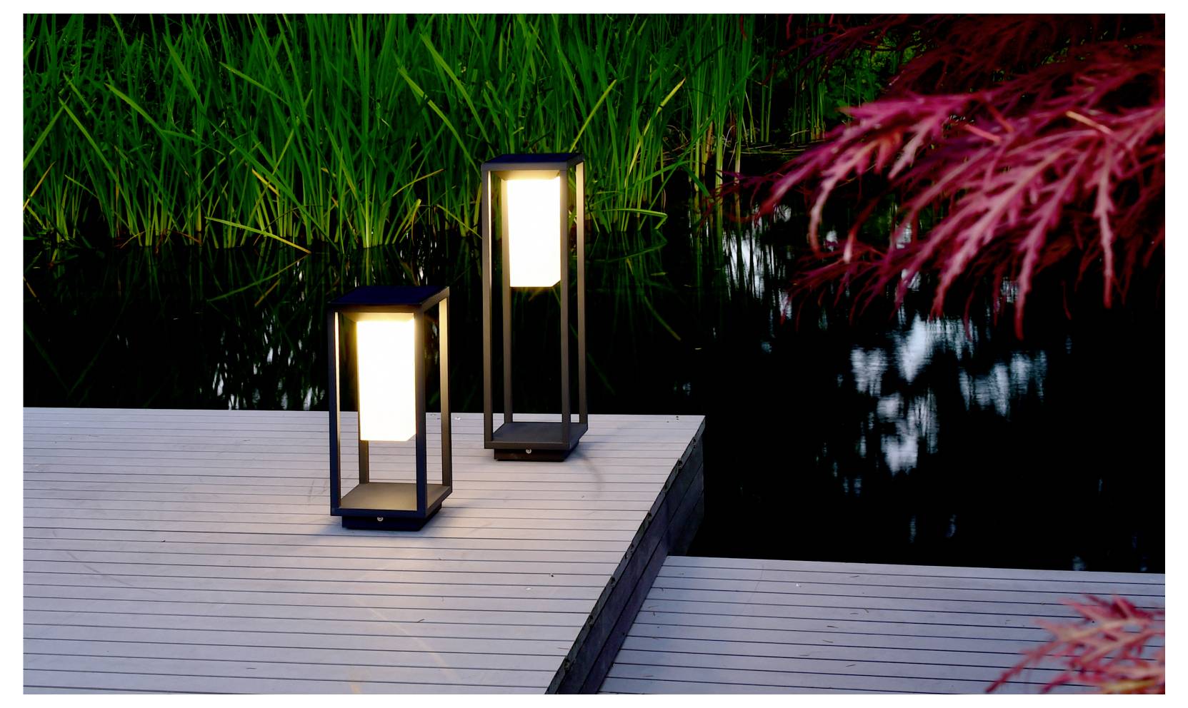 Due lanterne luminose su un ponte di legno accanto a uno stagno scuro, con canne verdi e foglie rosse che si riflettono nell'acqua.